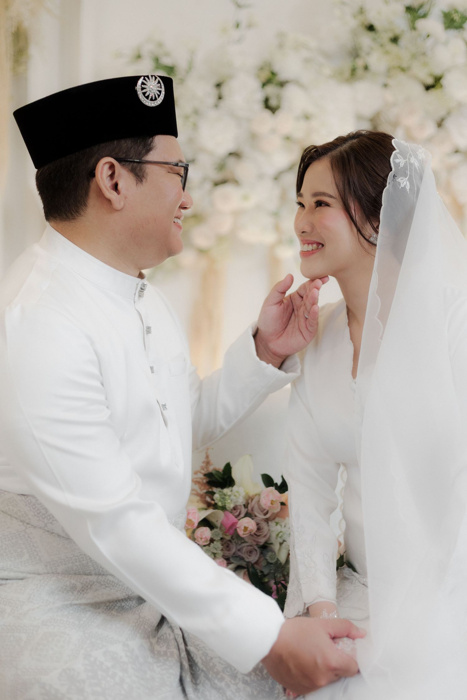A couple in traditional Malay wedding attire share a tender moment, the groom gently touching the bride's chin.