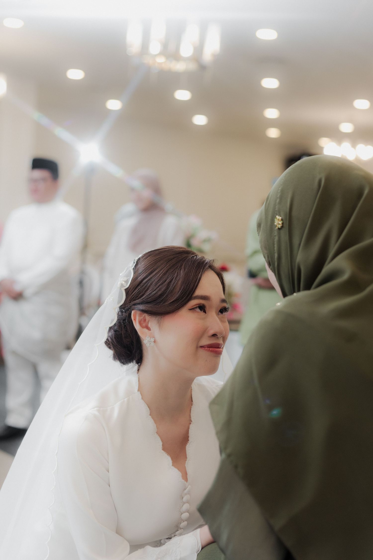 Bride in white dress smiling at a guest in green hijab during a traditional Malay wedding ceremony.