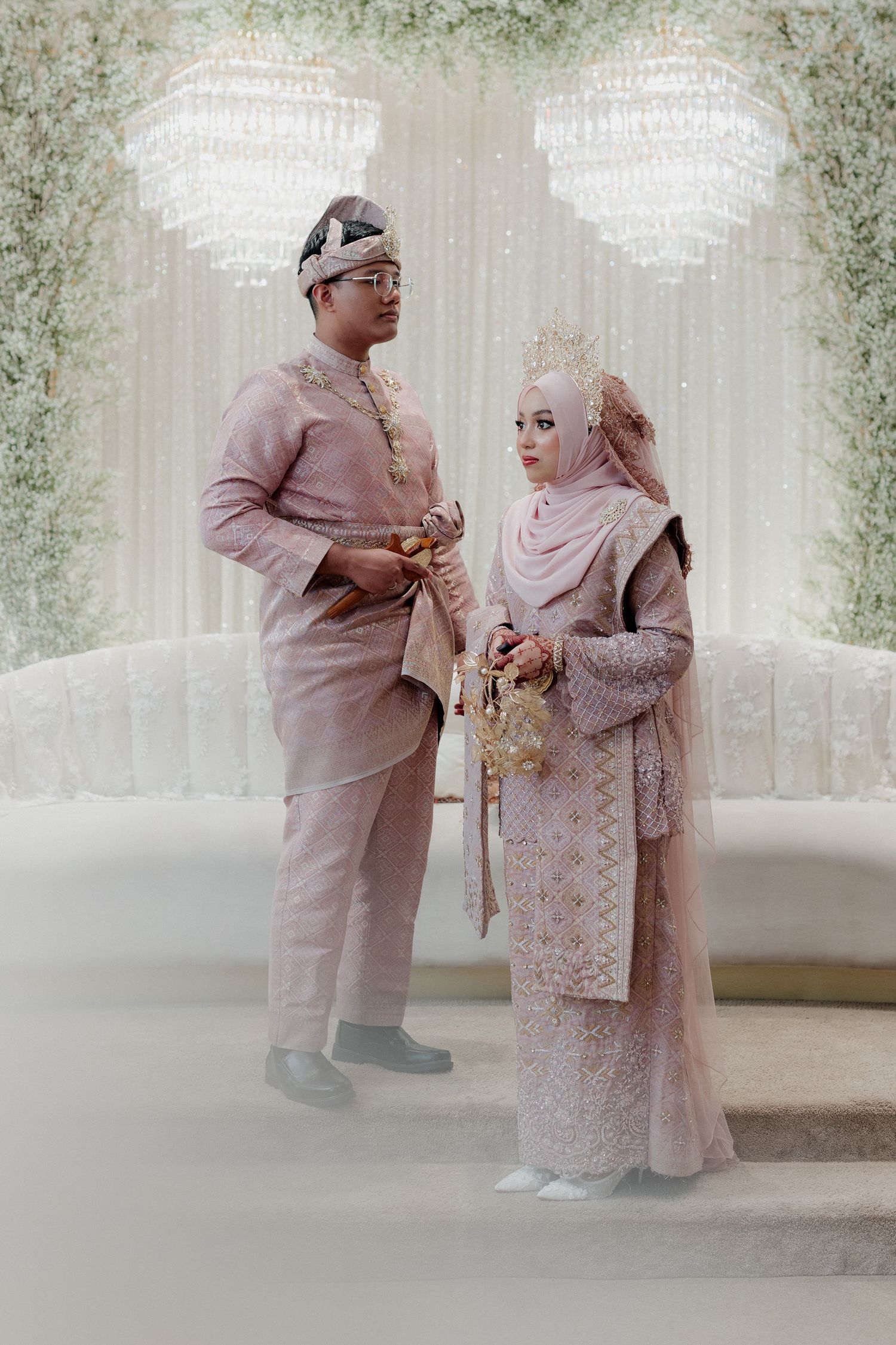 Muslim couple in matching blush pink traditional wedding attire standing elegantly at a decorated ceremony venue.