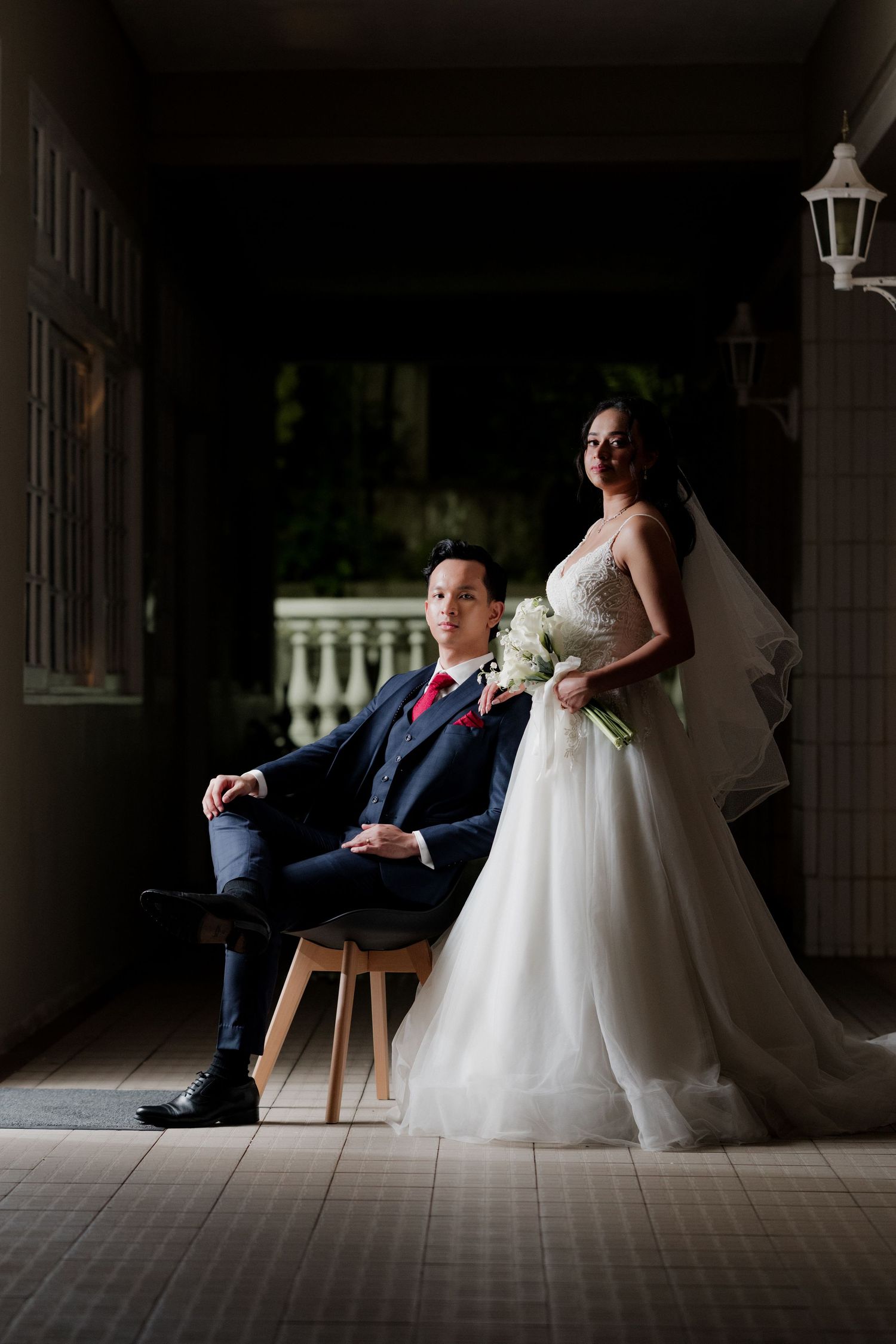 Bride in white gown holding bouquet stands beside groom seated in chair during elegant wedding portrait session.