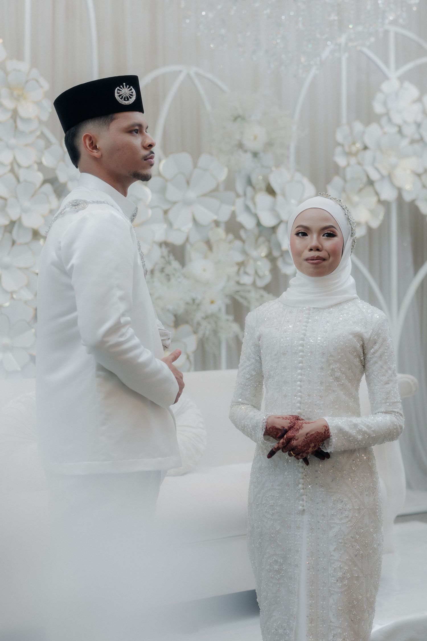 Malay couple in white traditional wedding attire posing before floral backdrop at elegant ceremony.