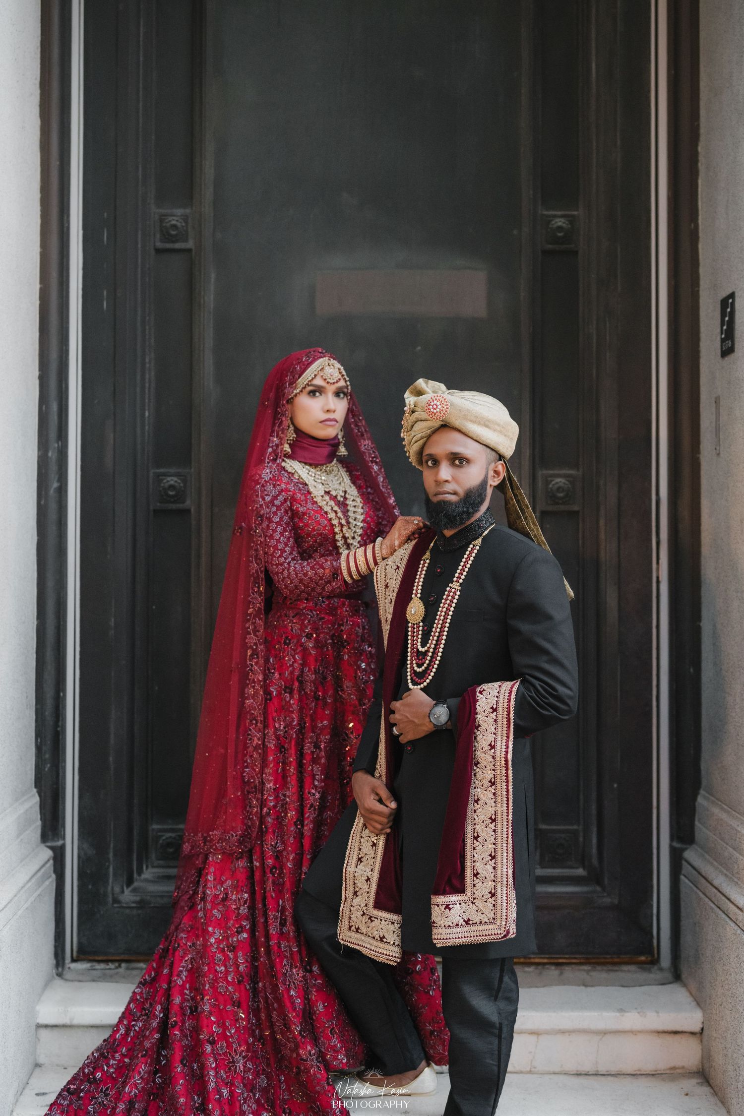 South Asian bride in red lehenga and groom in black sherwani with gold turban pose at ornate door.