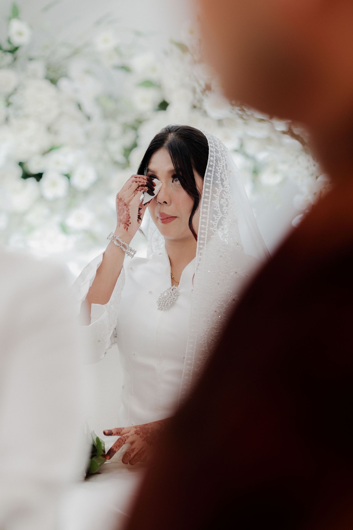 Emotional bride in white hijab and gown wiping tears during wedding ceremony with floral backdrop.