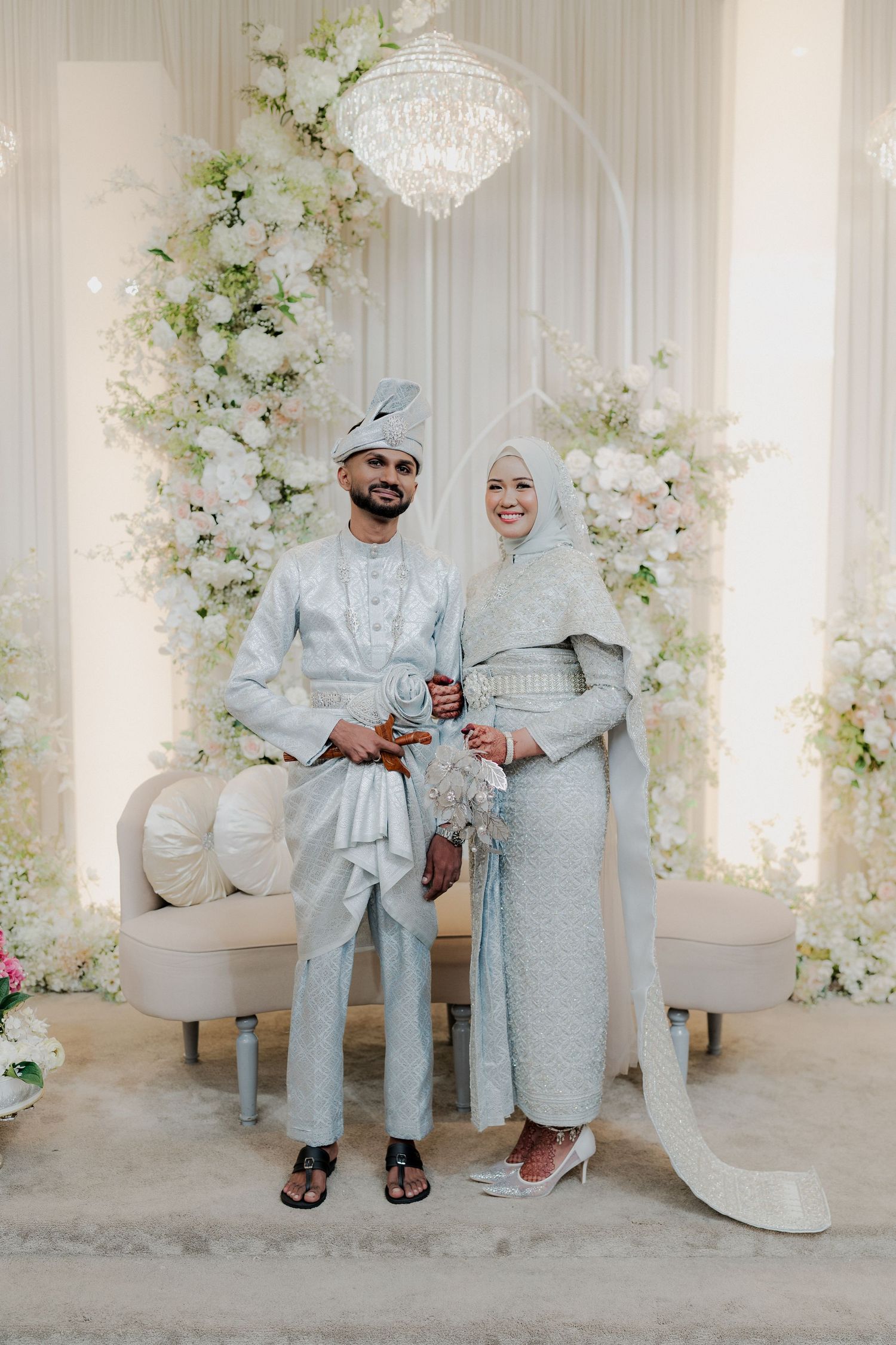 Indian and chinese couple in silver traditional wedding attire standing before floral arch with chandelier backdrop.