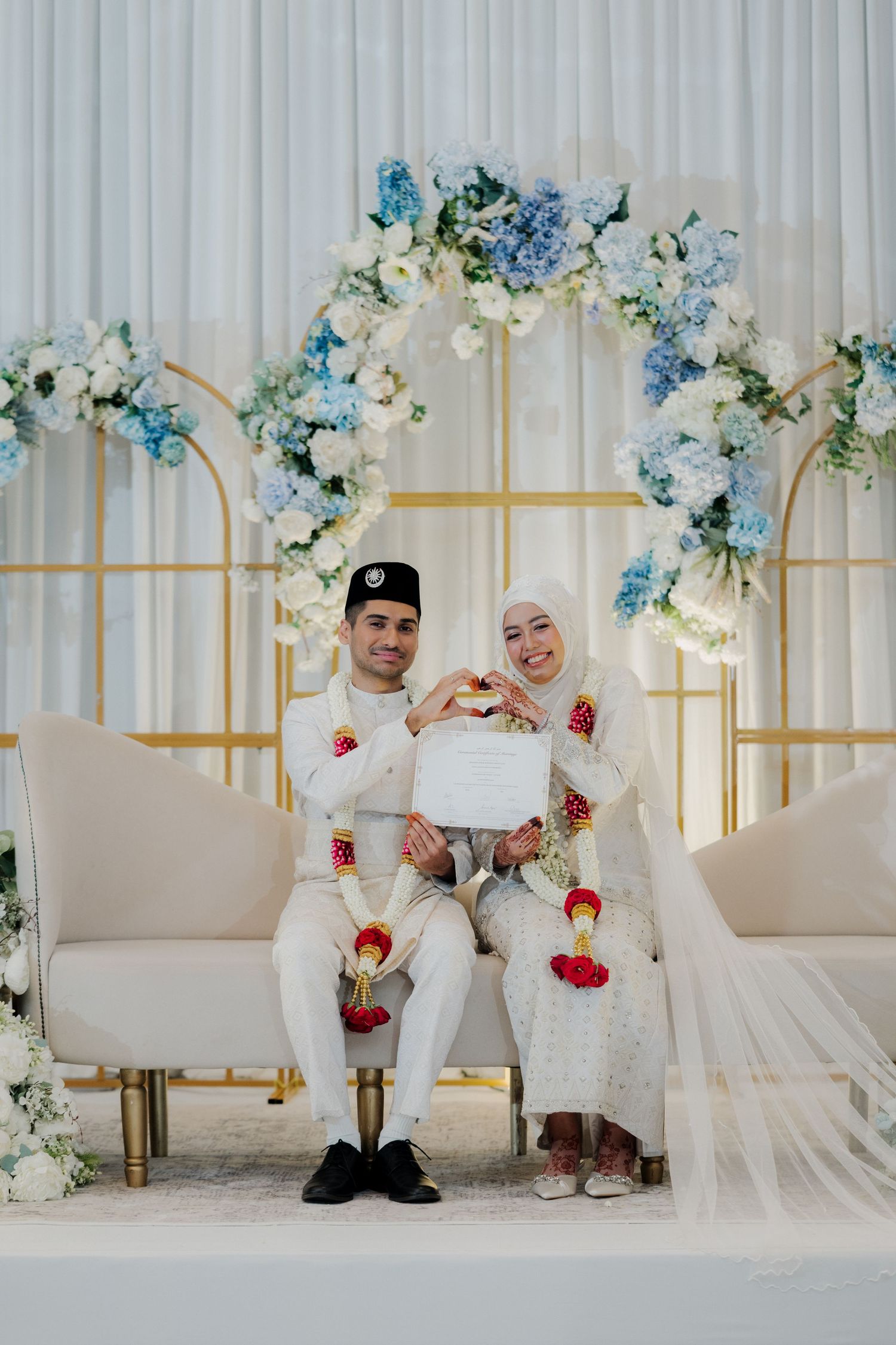 Muslim couple in traditional white wedding attire holding marriage certificate, seated before floral arch backdrop.