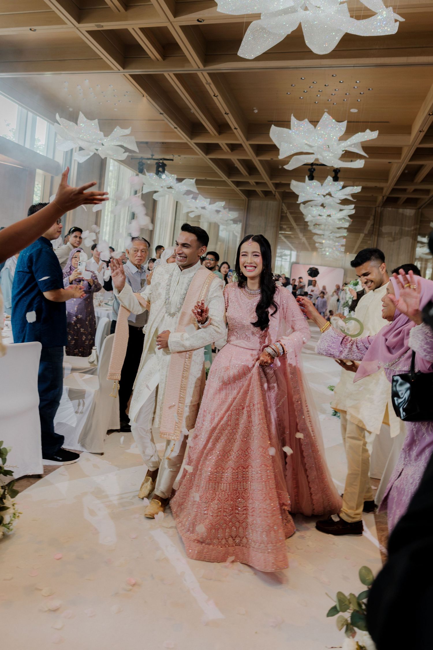 Bride in pink lehenga and groom in sherwani walk down aisle at elegant Indian wedding reception with floral decor.
