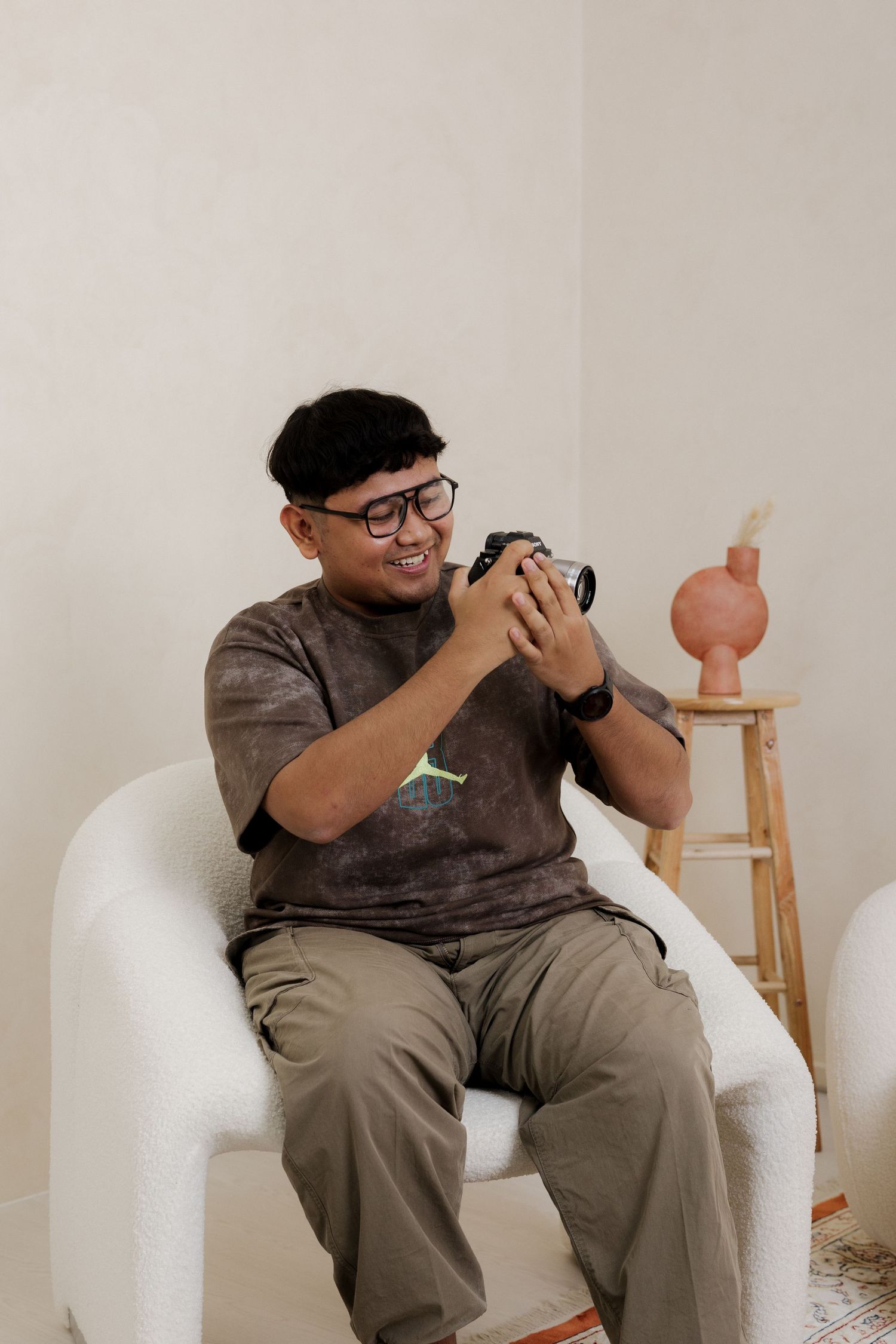 Young man in glasses and Jordan t-shirt smiles while reviewing photos on camera, seated in white armchair.