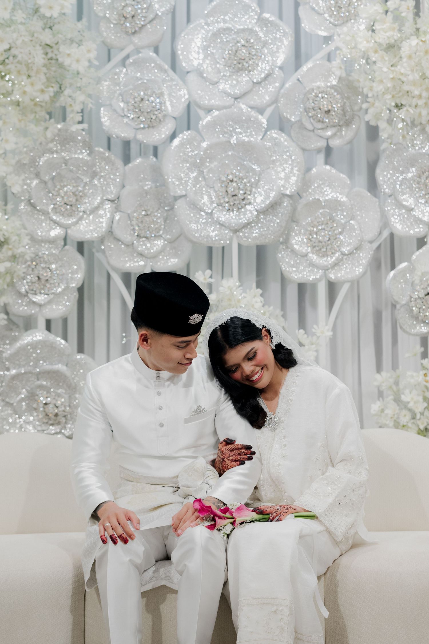 Malay couple in white traditional wedding attire sitting together in front of elegant white floral backdrop.