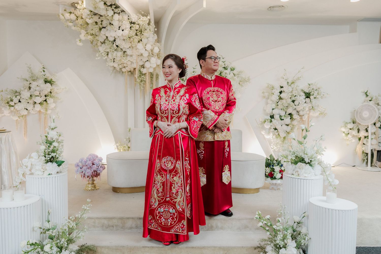 Interracial couple in traditional red Chinese wedding attire standing in an elegantly decorated white floral ceremony venue.