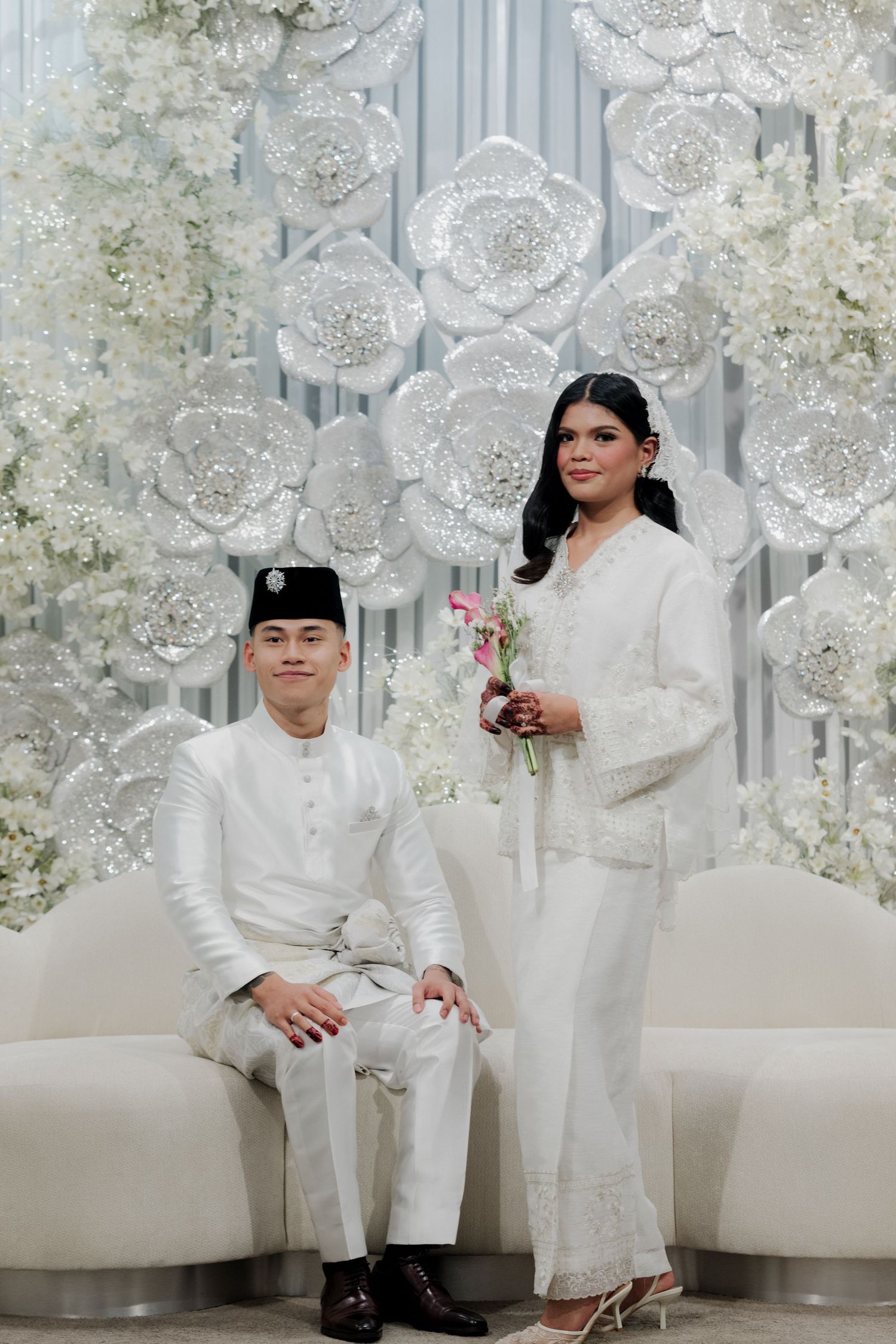 Malay couple in white traditional attire posing before elegant floral backdrop at a wedding ceremony.