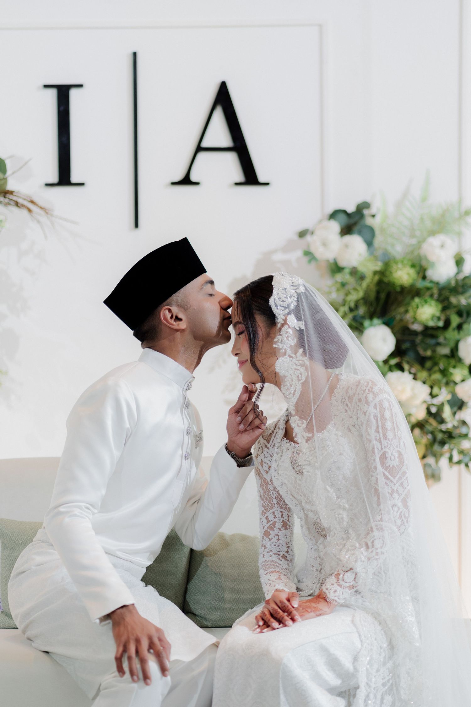 A Malay couple in white traditional wedding attire share a tender kiss at their elegant nikah ceremony.