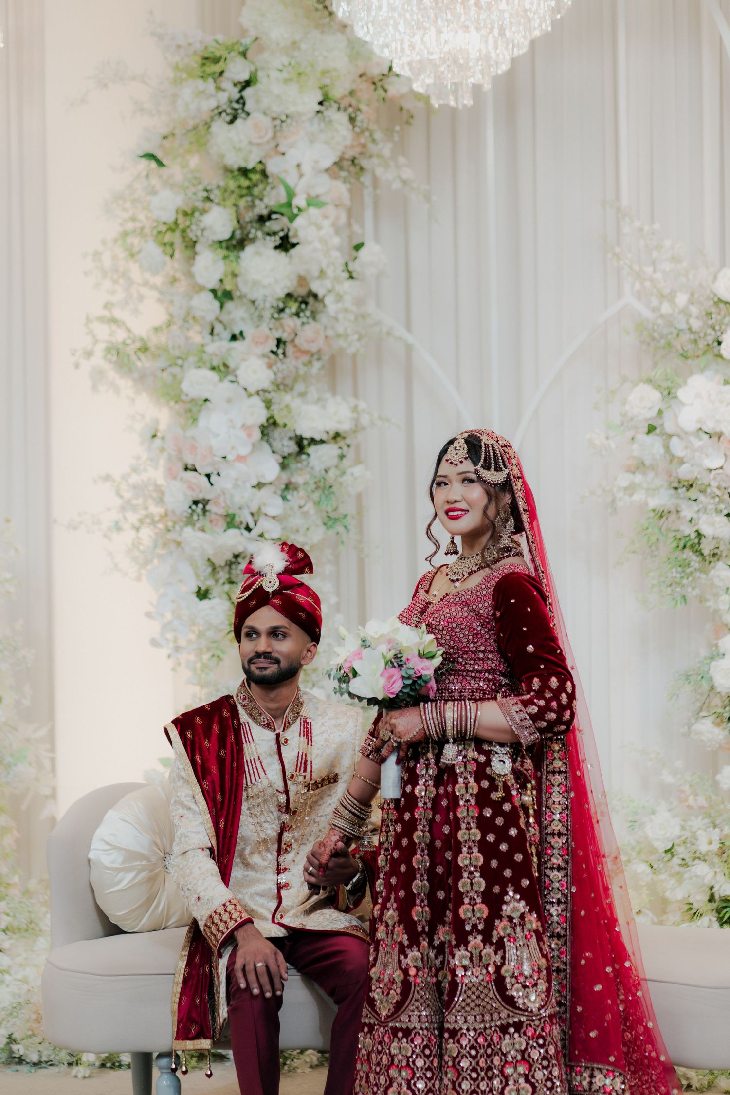 Indian bride in red lehenga and groom in sherwani pose before white floral backdrop at traditional wedding ceremony.