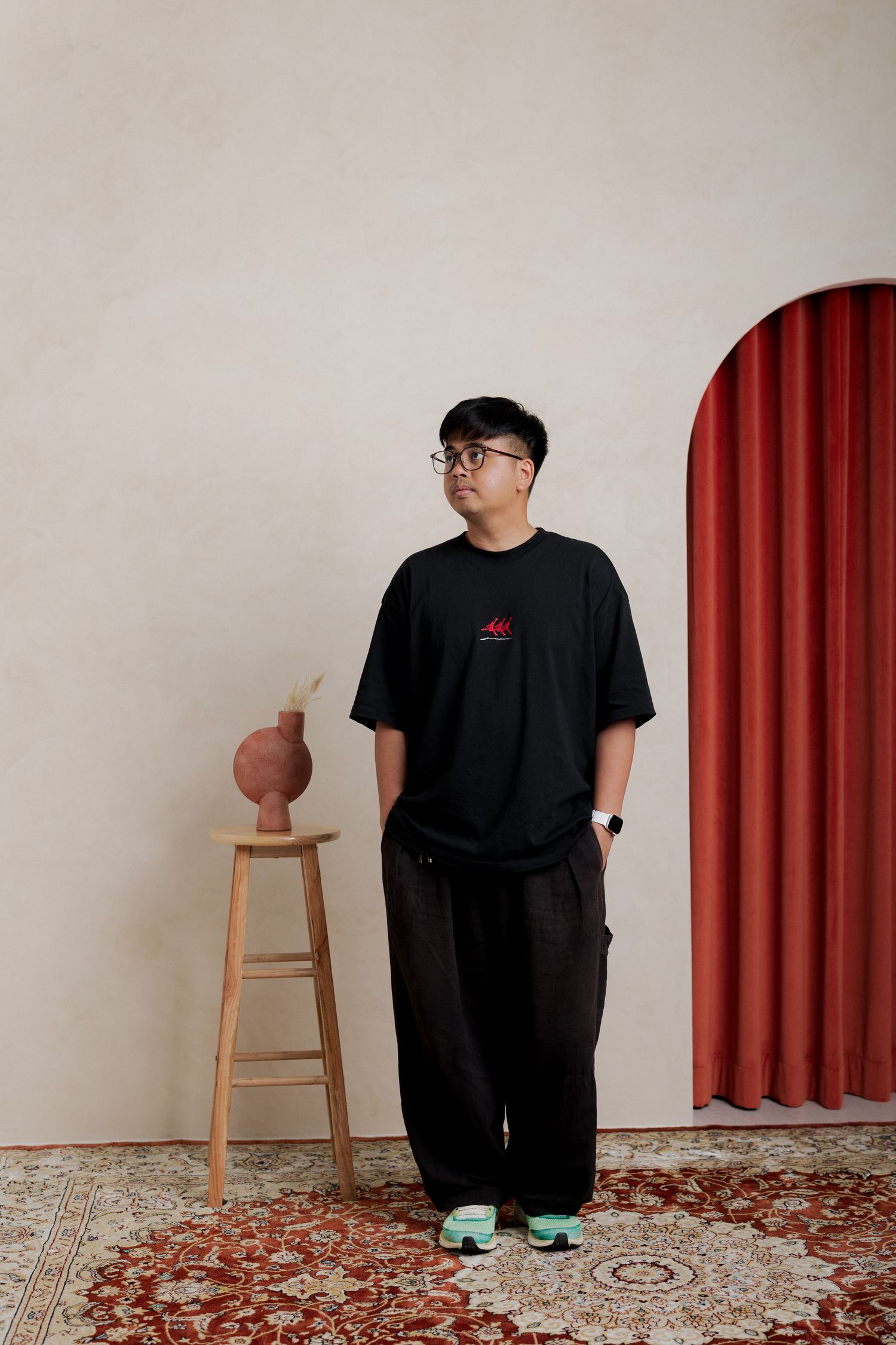 Young Asian man in black oversized t-shirt and dark trousers standing in minimalist studio with red curtain arch.