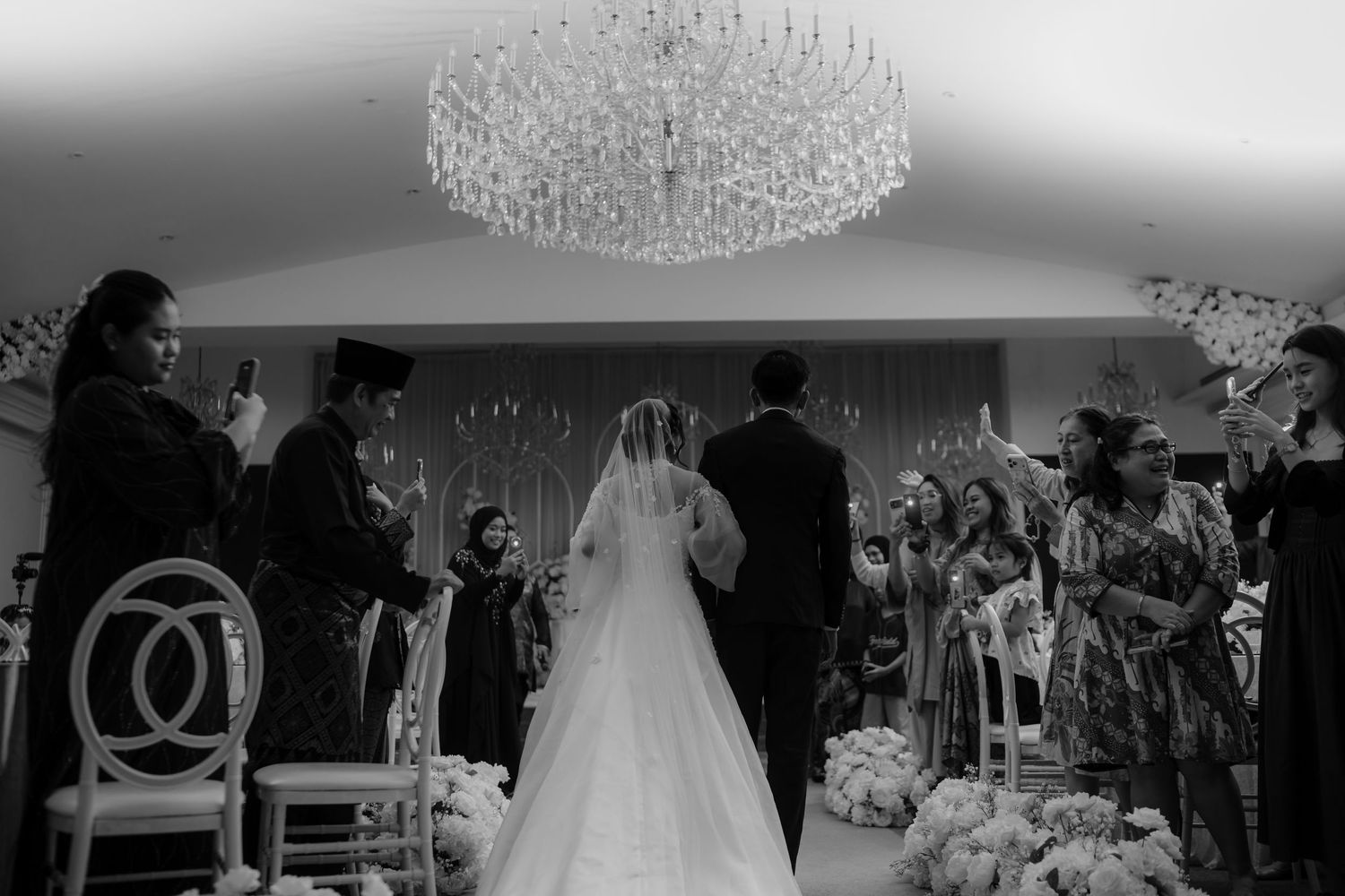 Black and white wedding ceremony photo with bride in gown under crystal chandelier, guests standing in elegant venue.