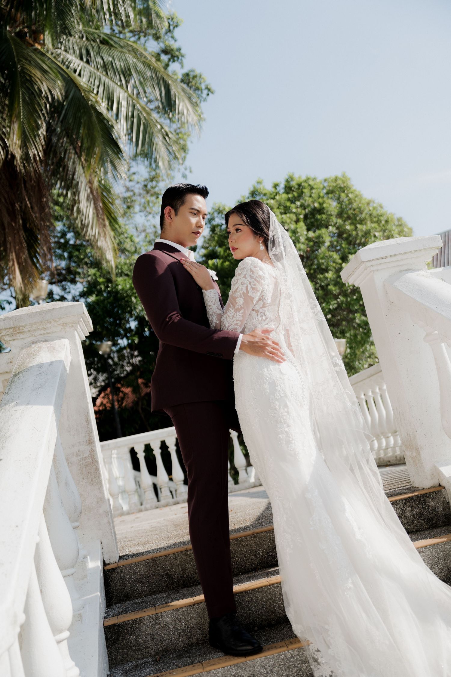 Bride and groom in elegant wedding attire pose on white balustrade steps surrounded by tropical greenery.