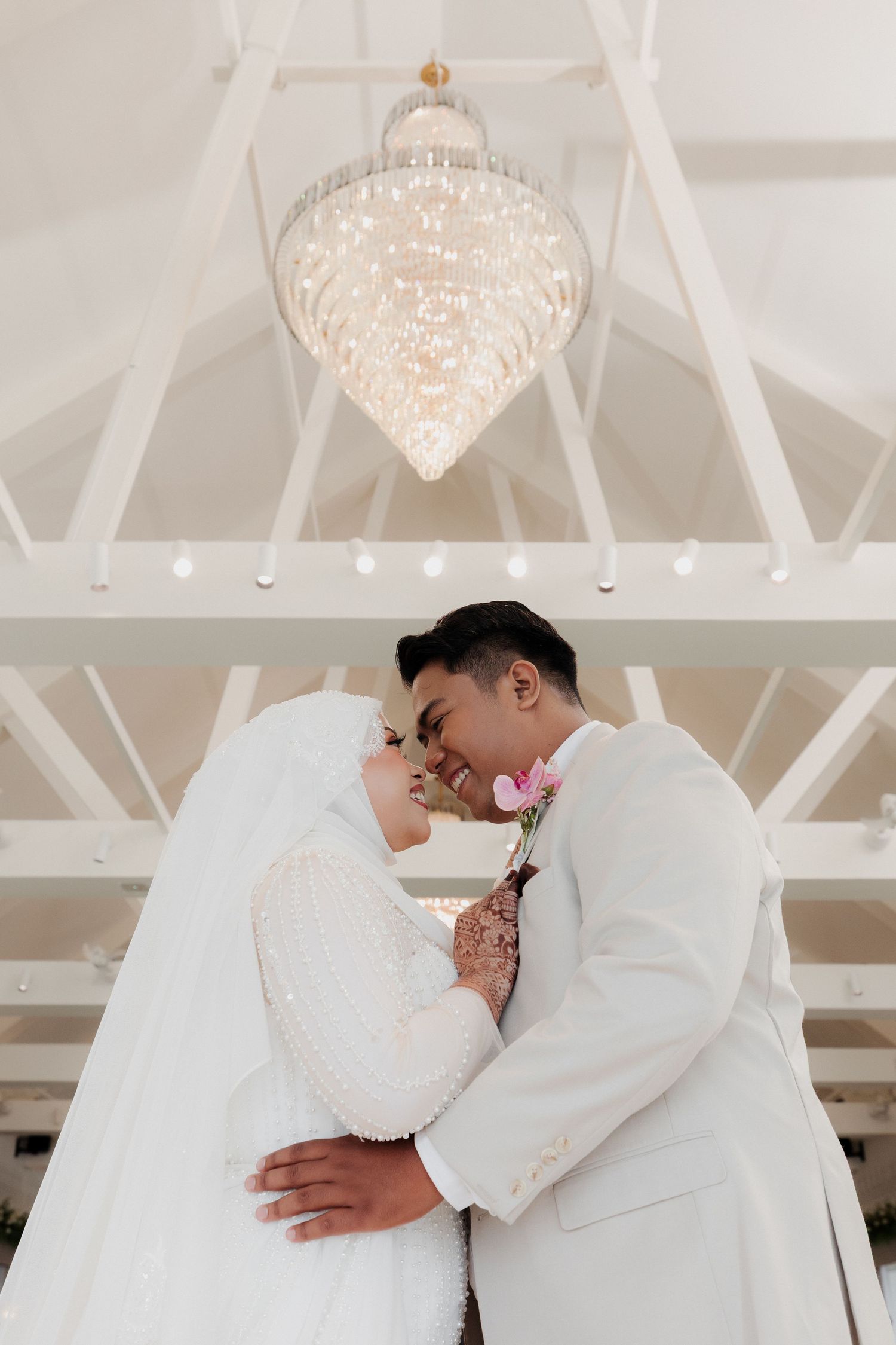 A couple in white wedding attire share a romantic forehead touch beneath an elegant chandelier in a bright venue.