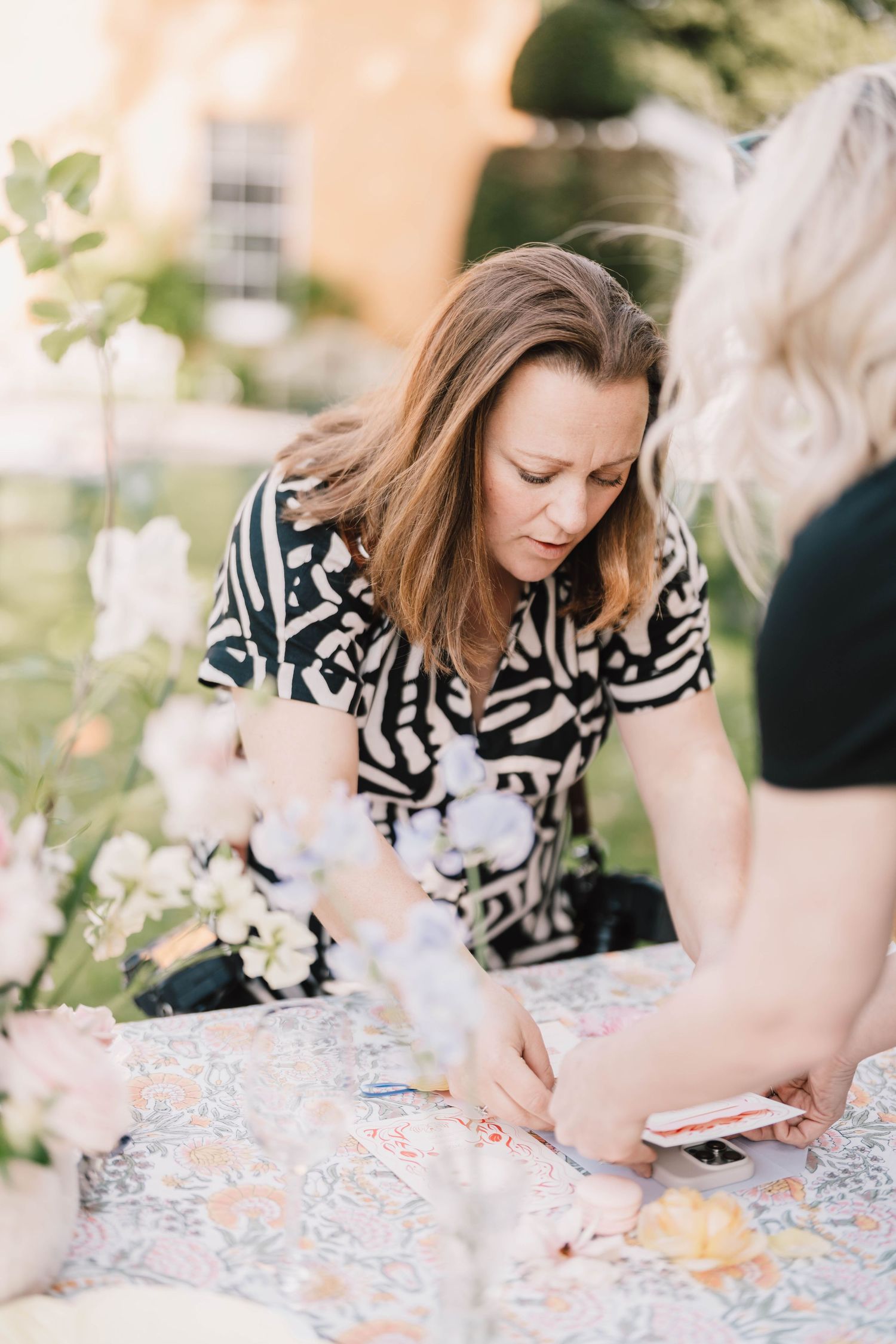 Woman in black and white patterned dress working on craft project at outdoor table with floral decorations.