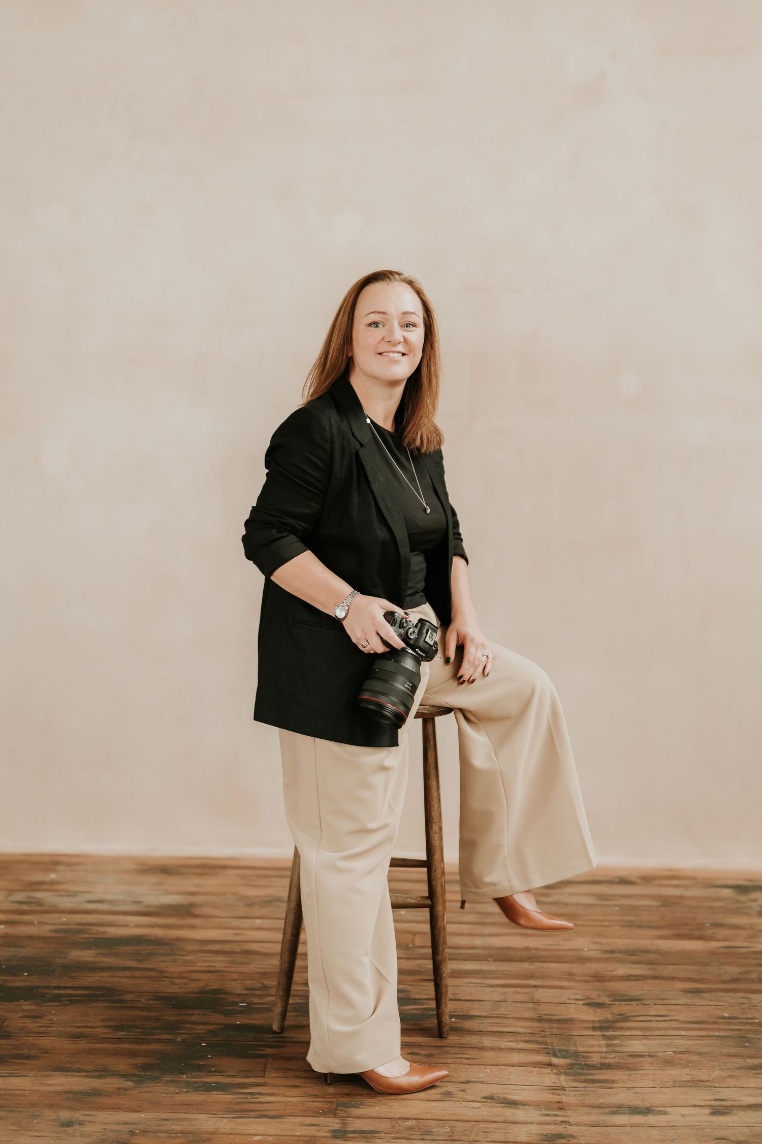 A Sophie Ruck in a black blazer and beige pants holds a camera while posing beside a stool.