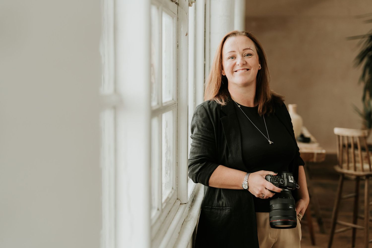 Smiling Sophie Ruck holding a DSLR camera standing by a bright window in a light-filled studio space.