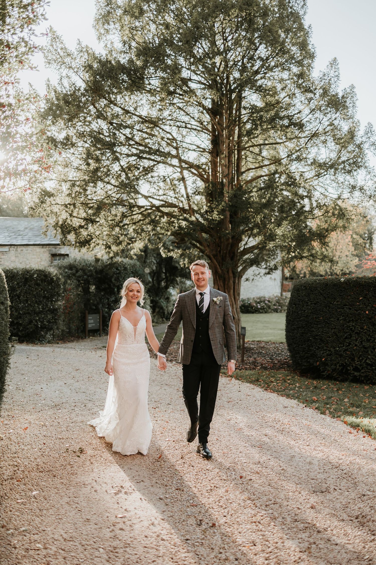 Newlyweds walking hand in hand down a tree-lined gravel path at sunset.