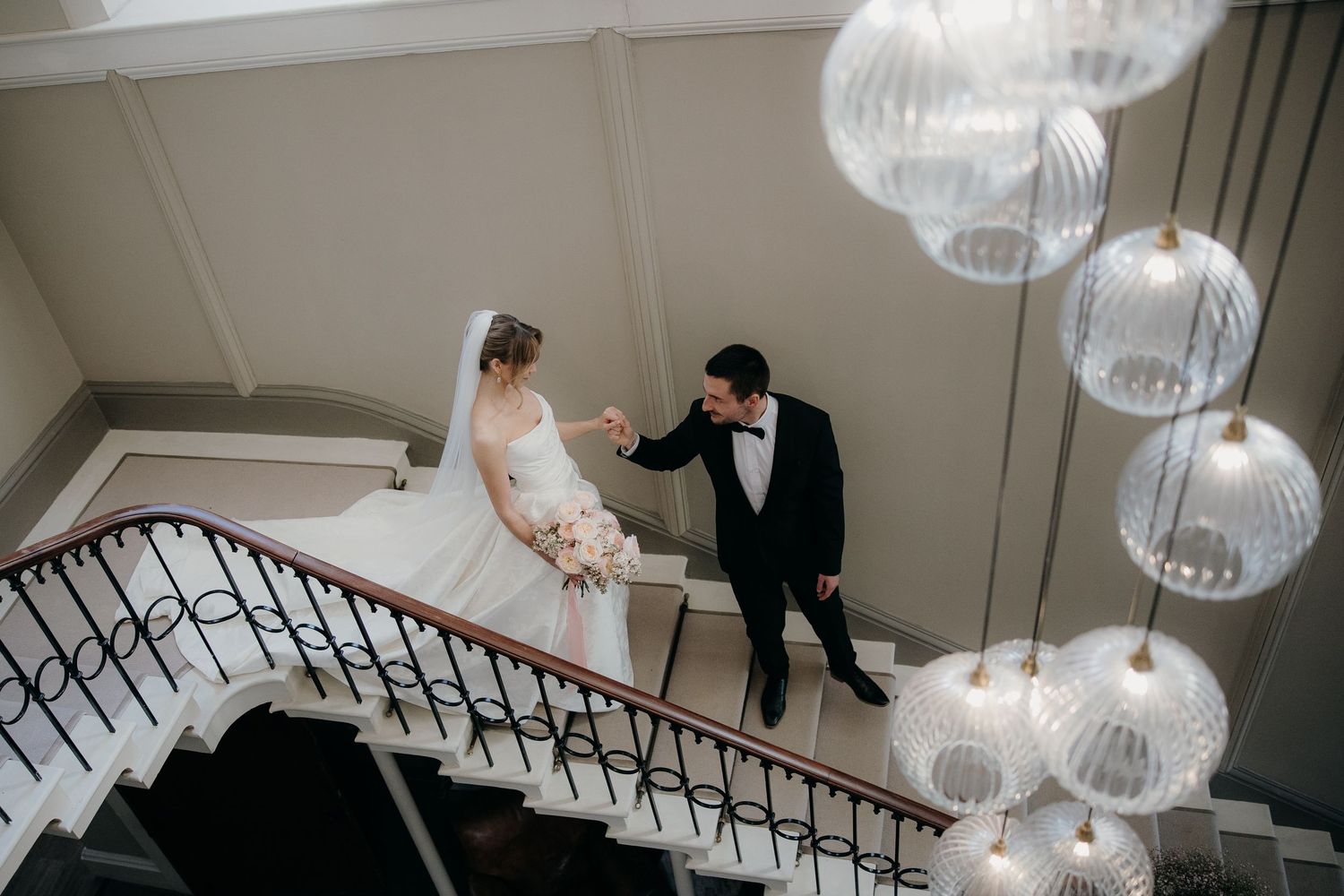 Bride and groom hold hands on elegant staircase with glass globe chandeliers at luxury wedding venue Saltmarshe hall.