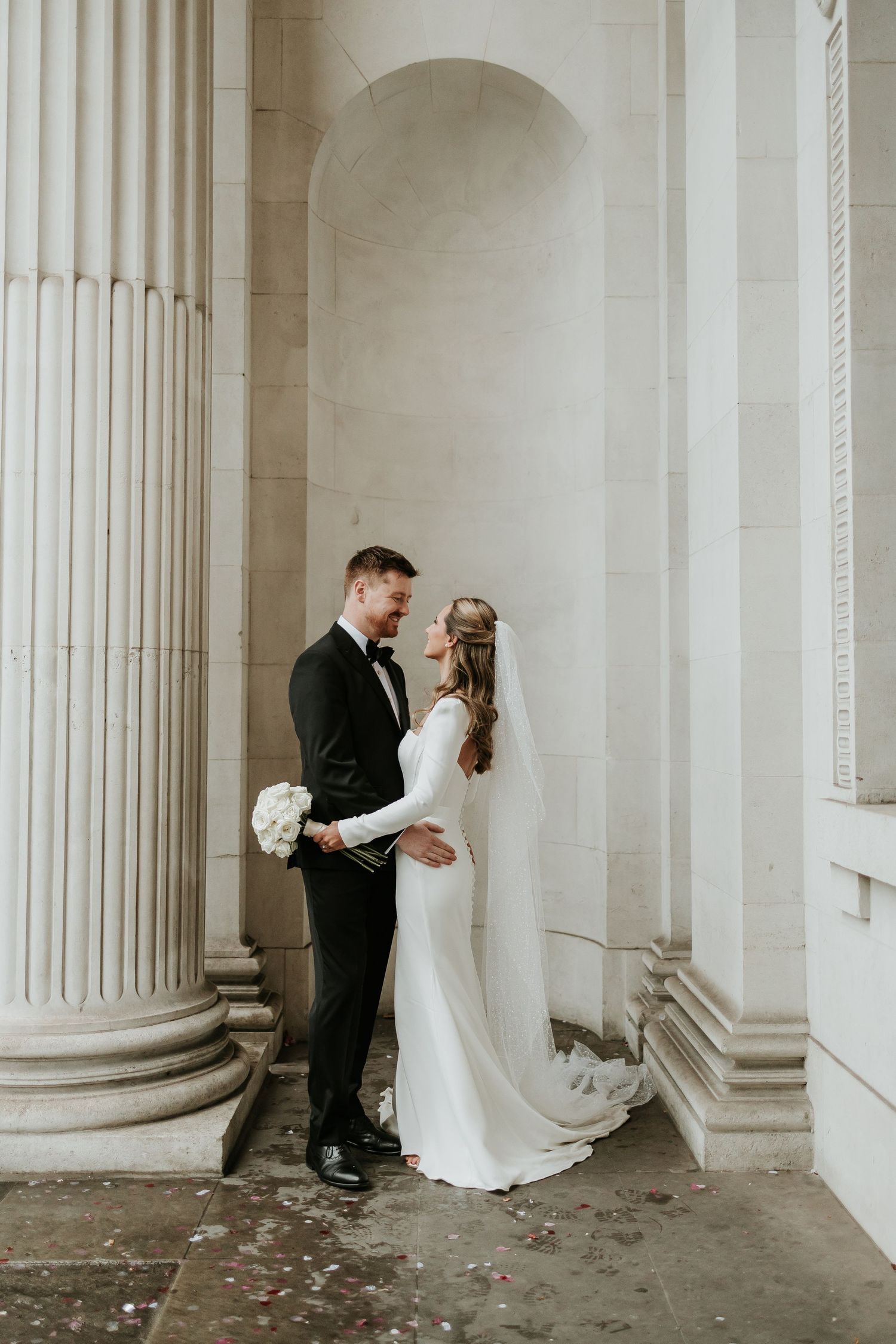 A wedding portrait of a couple in wedding attire embraces between elegant white columns of Marylebone Town Hall London.