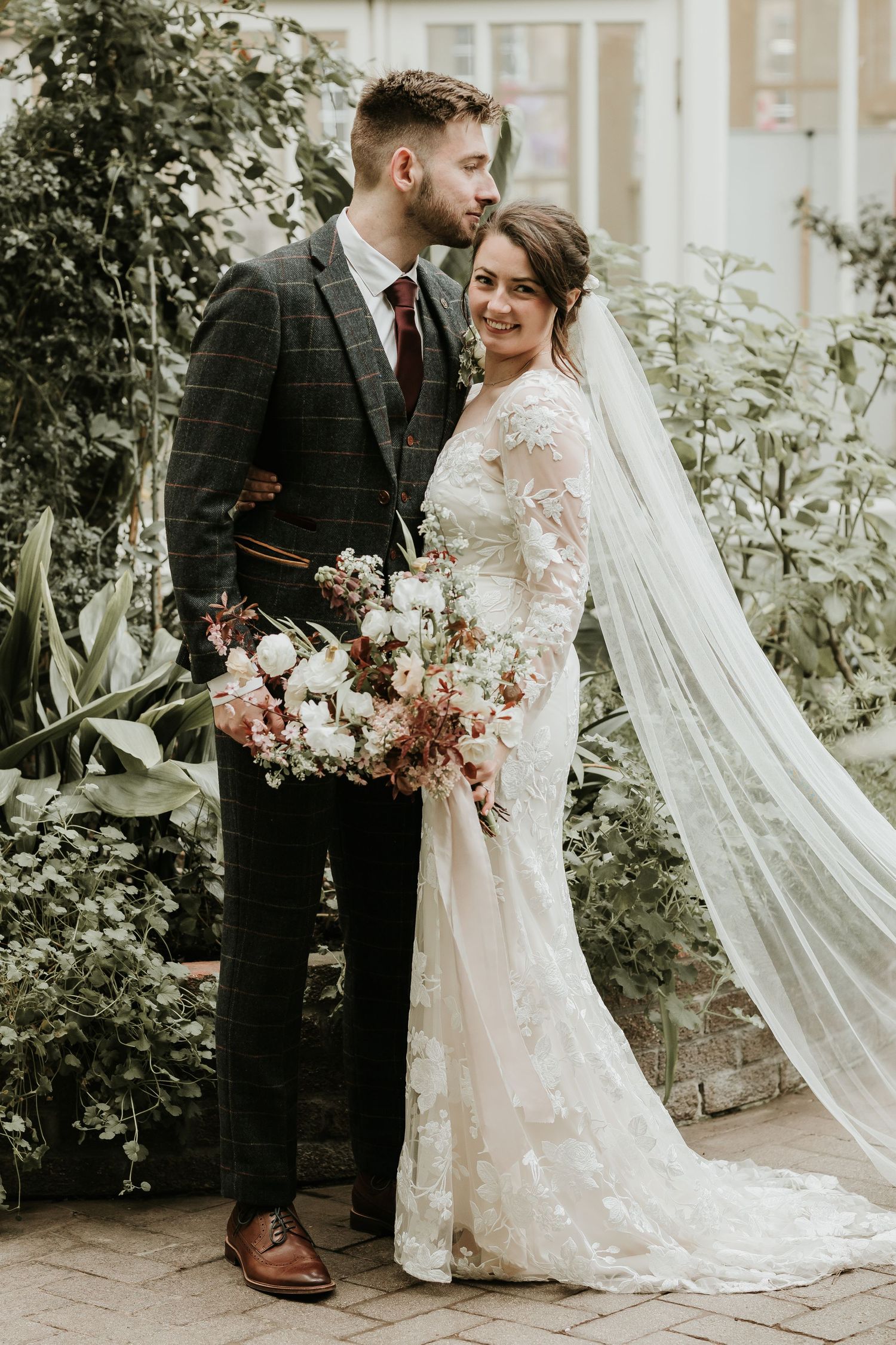 Wedding couple poses in garden with bride wearing lace gown and long veil. Palm house wedding ceremony