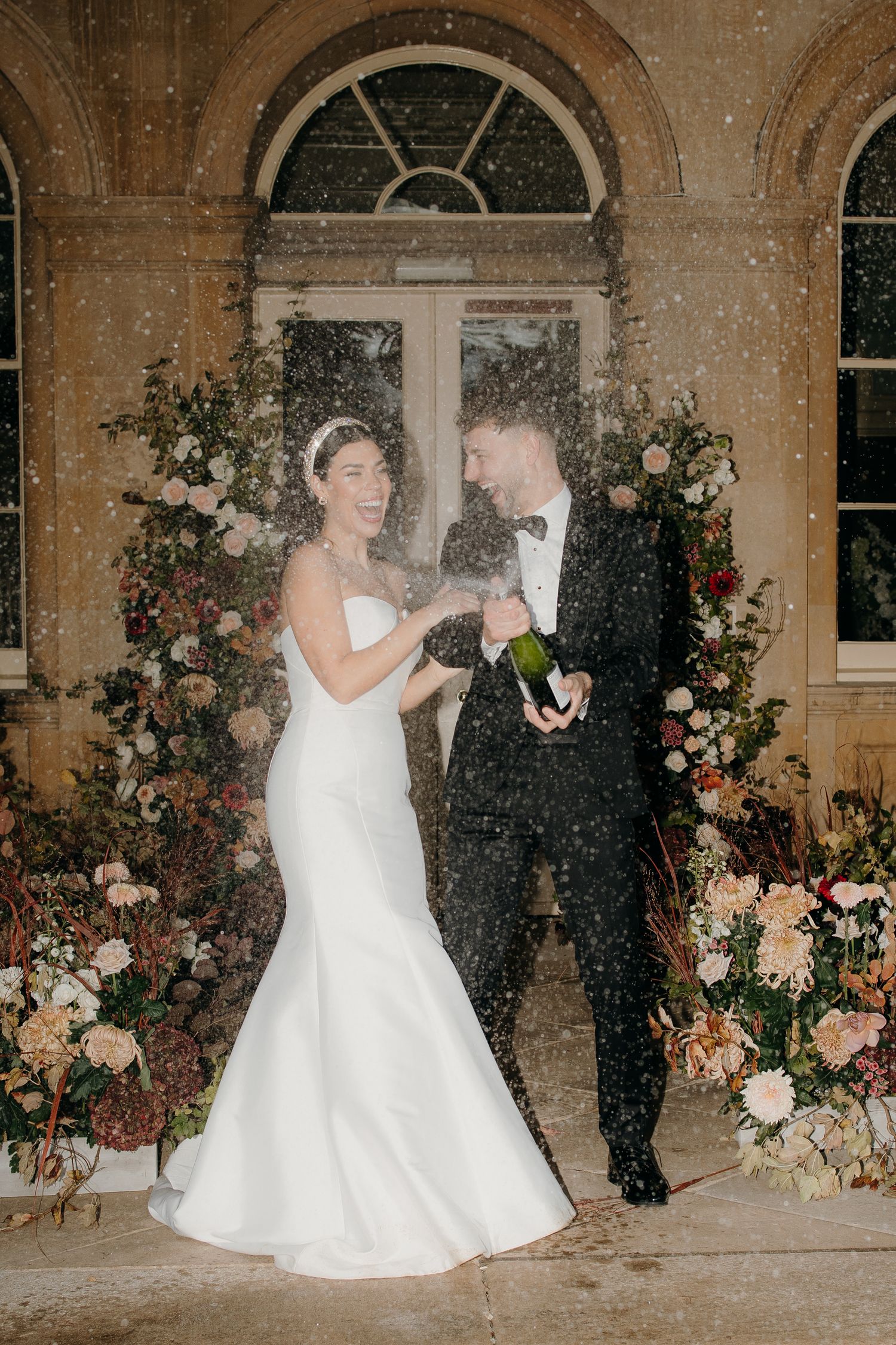Bride and groom popping champagne bottle outdoors at night, surrounded by lush floral arrangements at elegant wedding venue Prestwold hall.