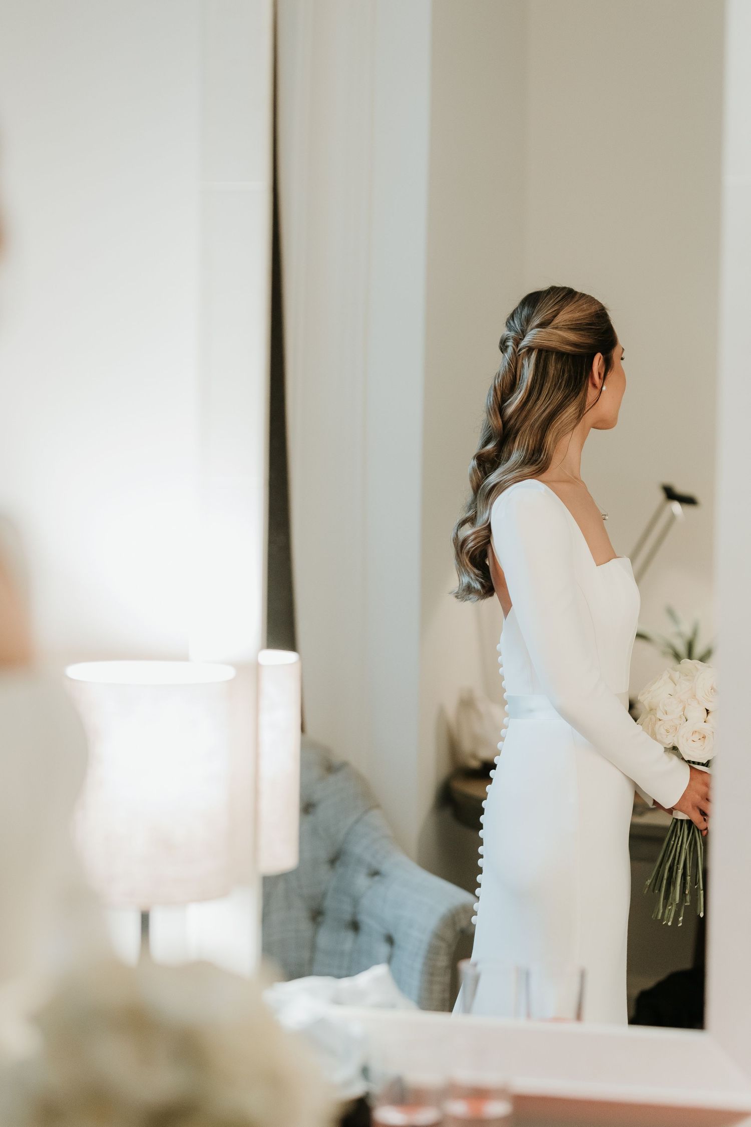 Bride in white long-sleeve wedding dress holds bouquet, viewed from behind in mirror reflection.