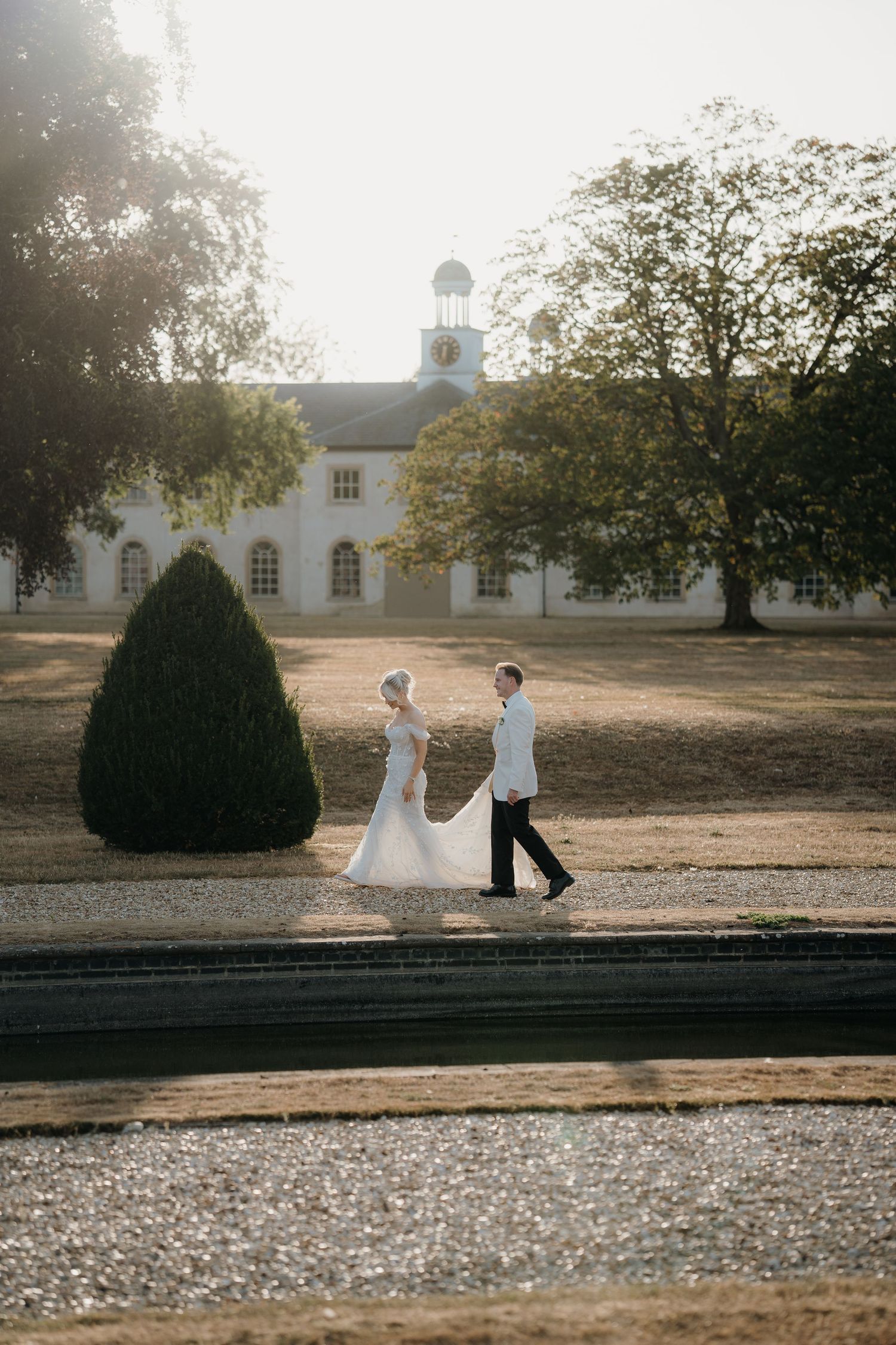 Bride in a flowing white gown walks hand in hand with her groom across a golden lawn toward a grand estate with a clock tower.