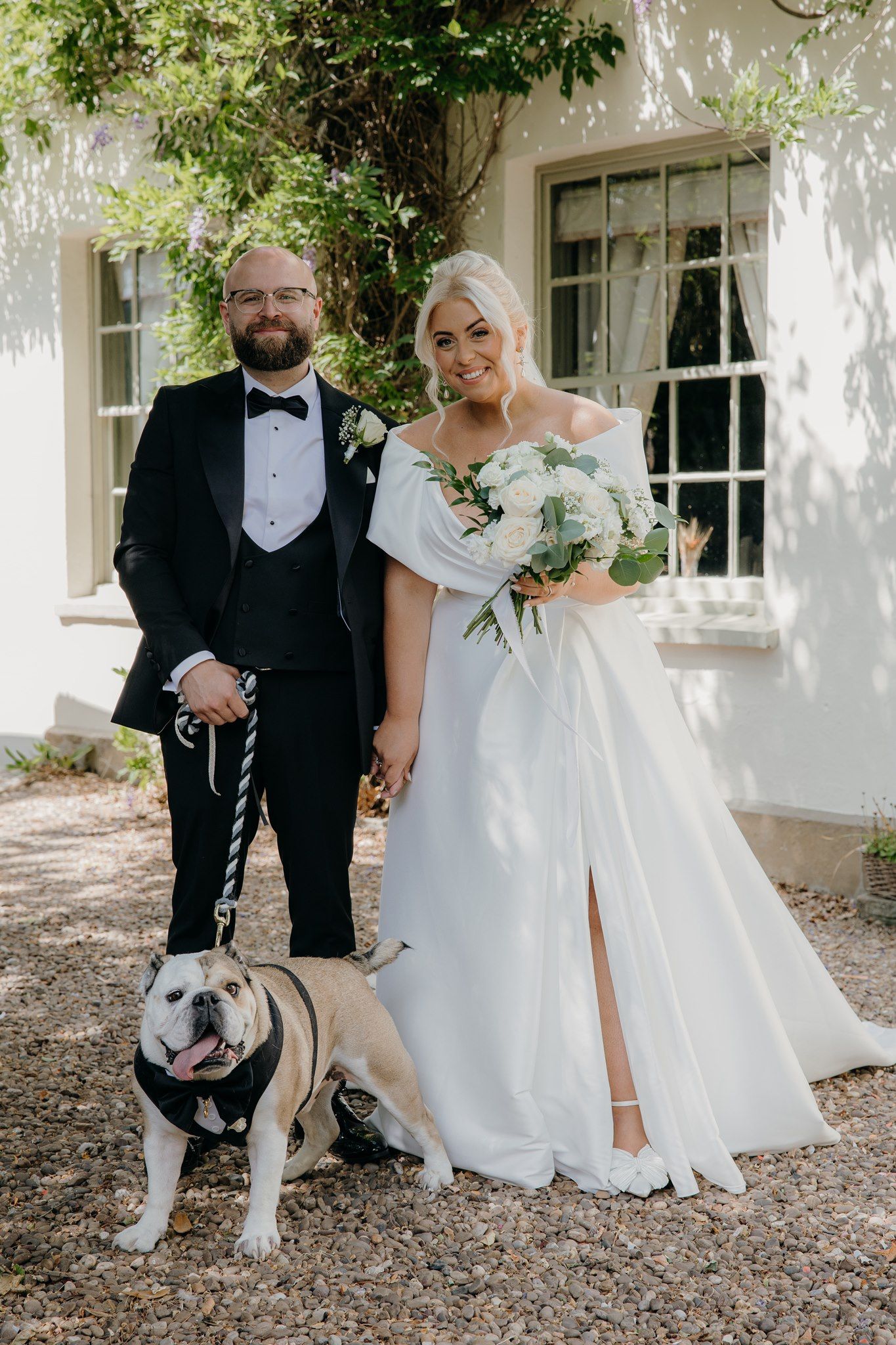 Bride in a white satin gown holding a bouquet and groom in a black tuxedo hold hands beside their English bulldog outside a white house.