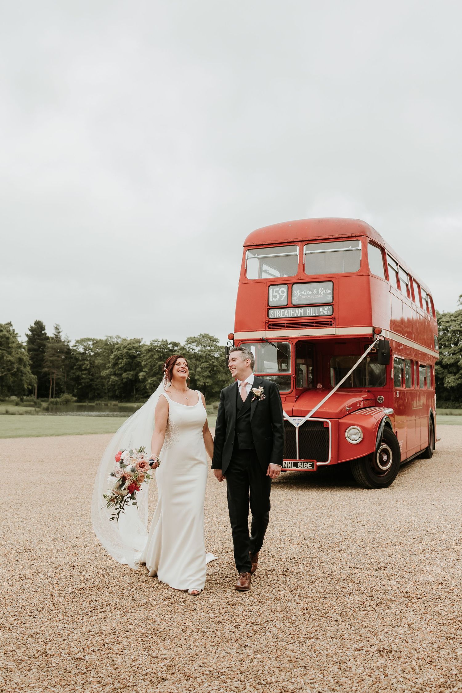 Bride and groom holding hands while walking past classic red London bus on overcast day at Stubton hall.