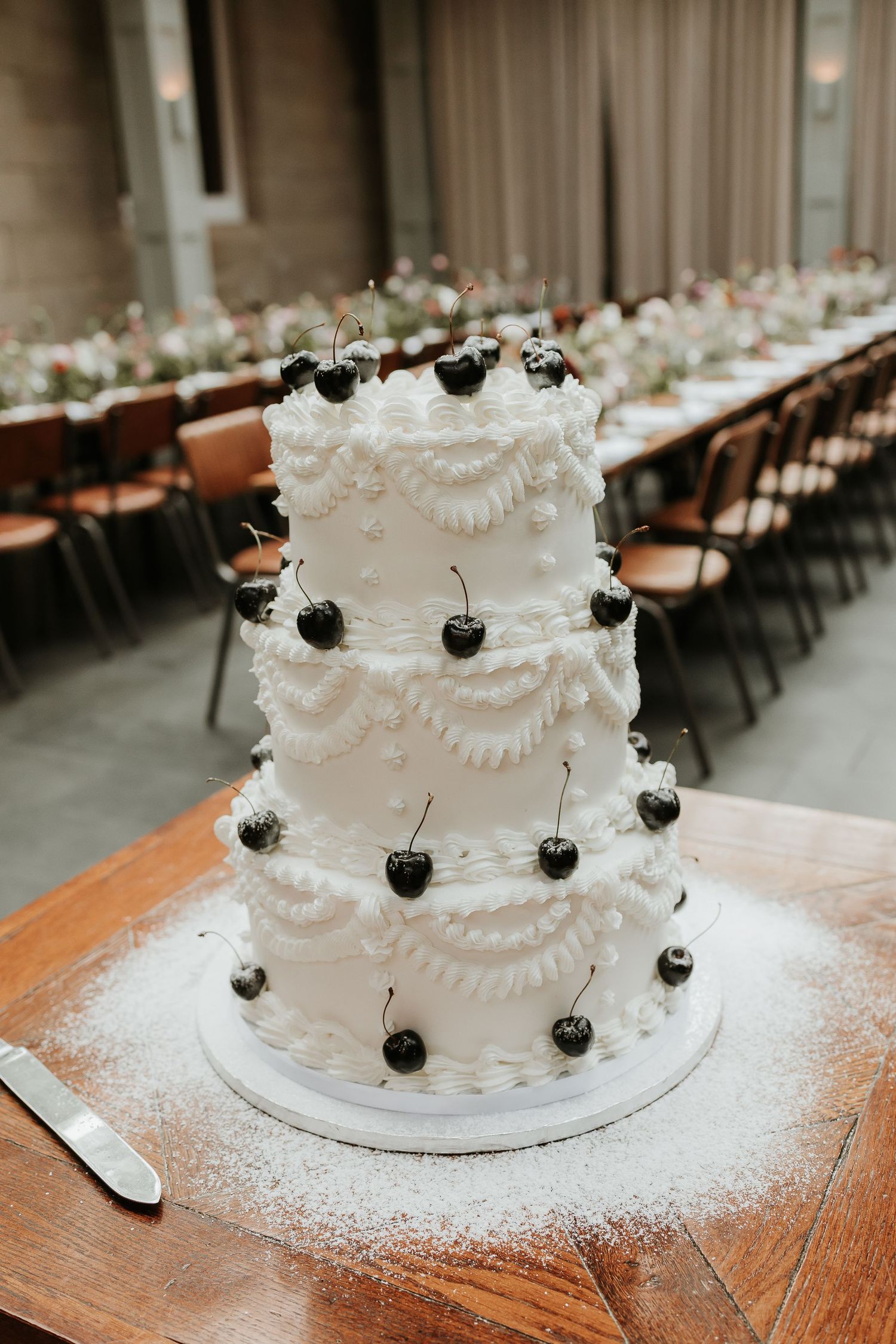 Three-tier white wedding cake decorated with fresh black cherries and piped frosting, displayed on wooden table in elegant reception hall.