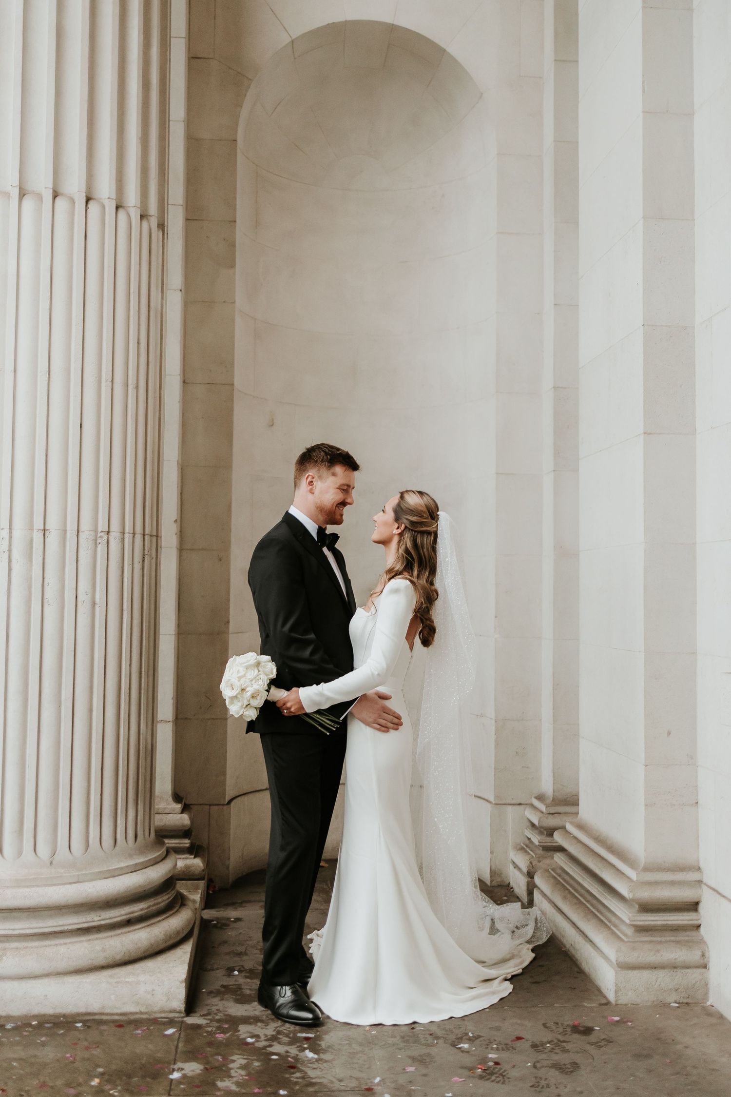Elegant wedding couple poses between classical stone columns in formal attire at Marylebone town hall. London