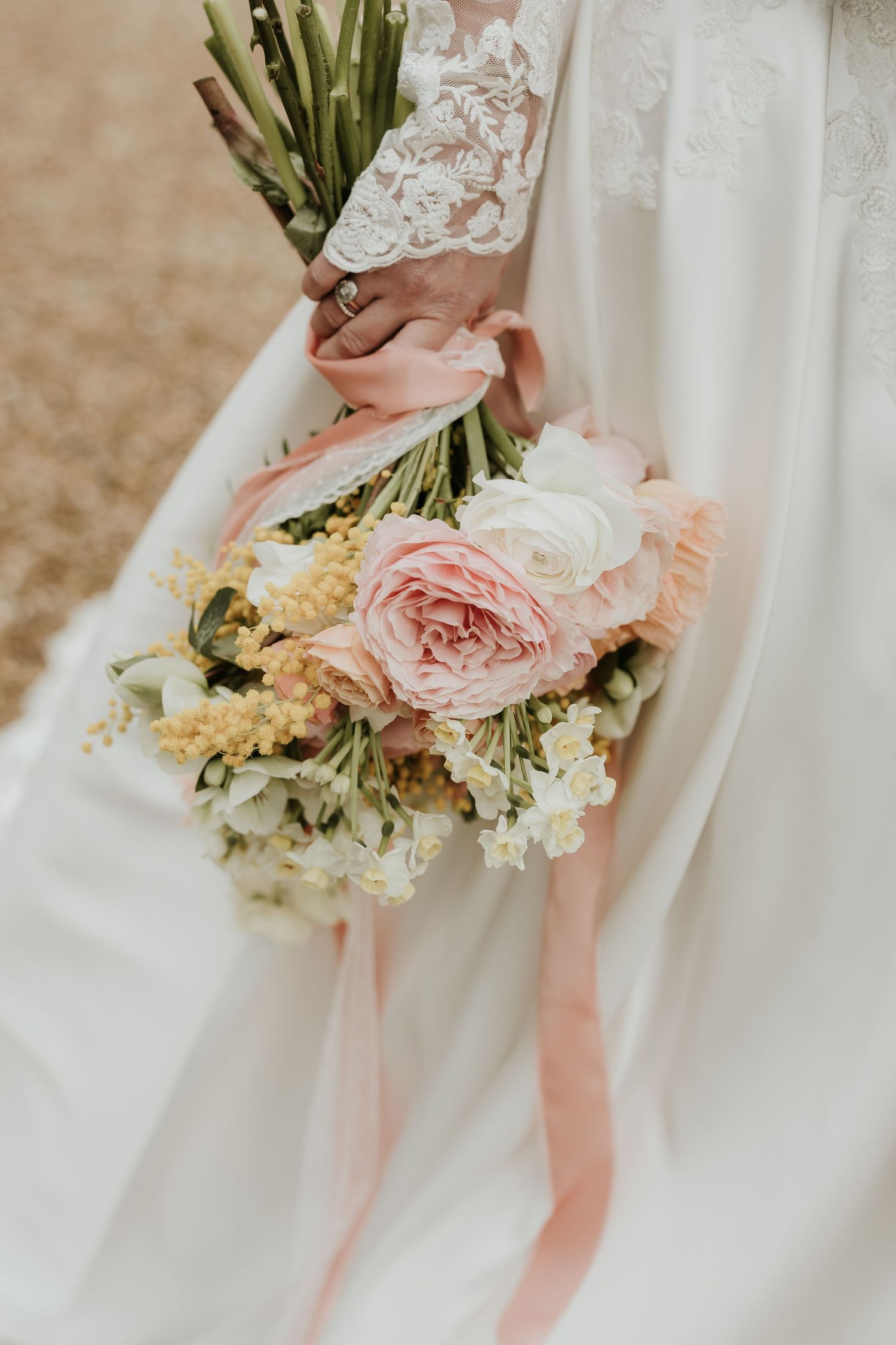 Bride in a white lace gown holding a romantic bouquet of blush pink roses, white blooms, and yellow mimosa tied with peach ribbons.