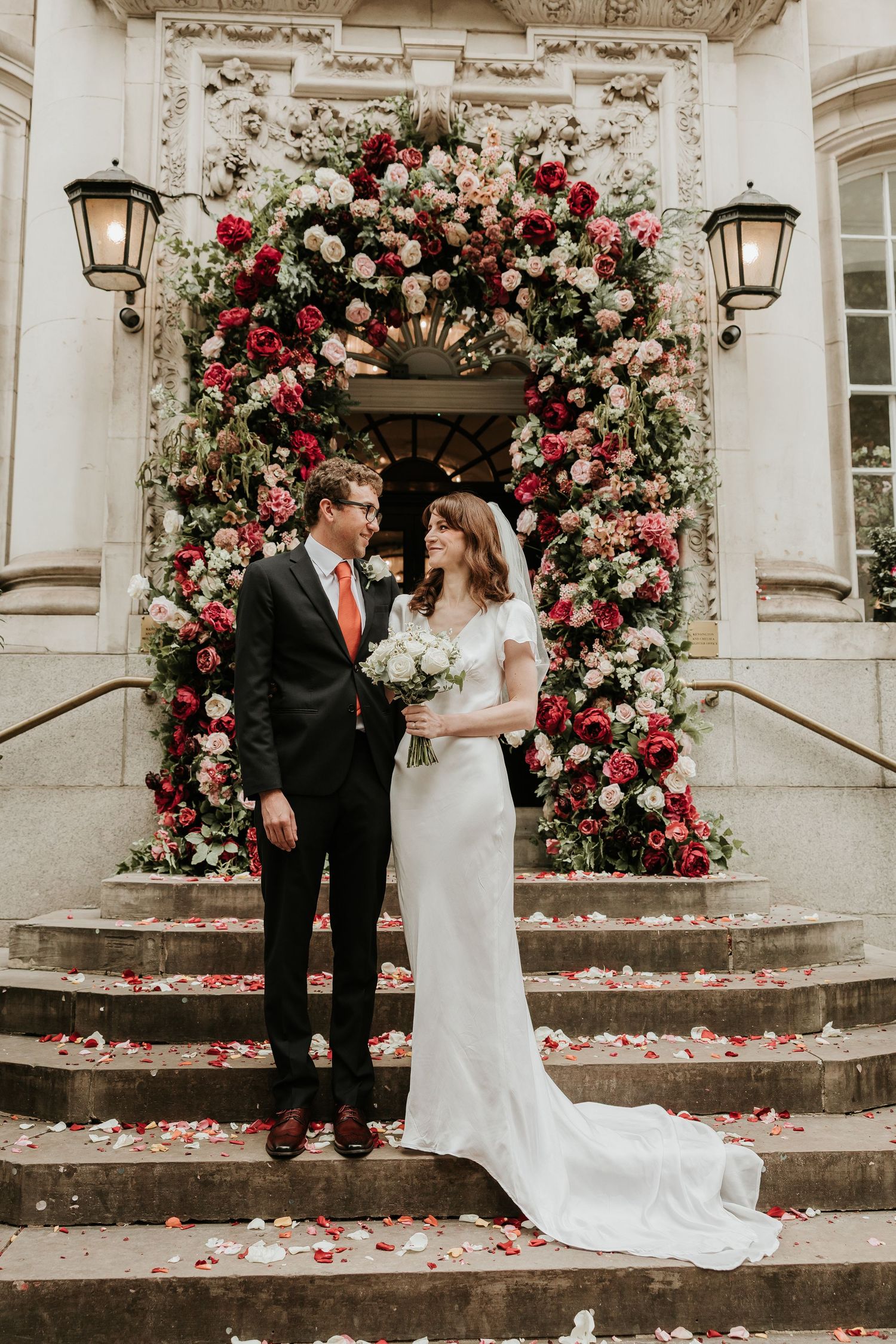 Couple stands on stone steps adorned with lush pink and red rose floral arrangements at an elegant venue entrance.