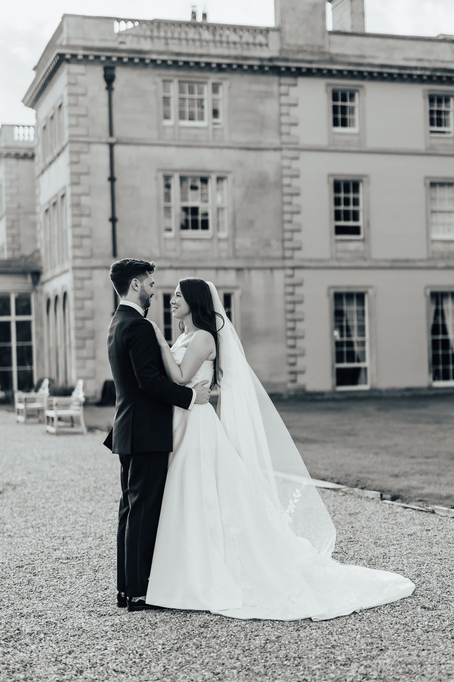 Bride in a white gown and long veil embraces her groom in a dark suit on a gravel courtyard before a grand stone manor house.