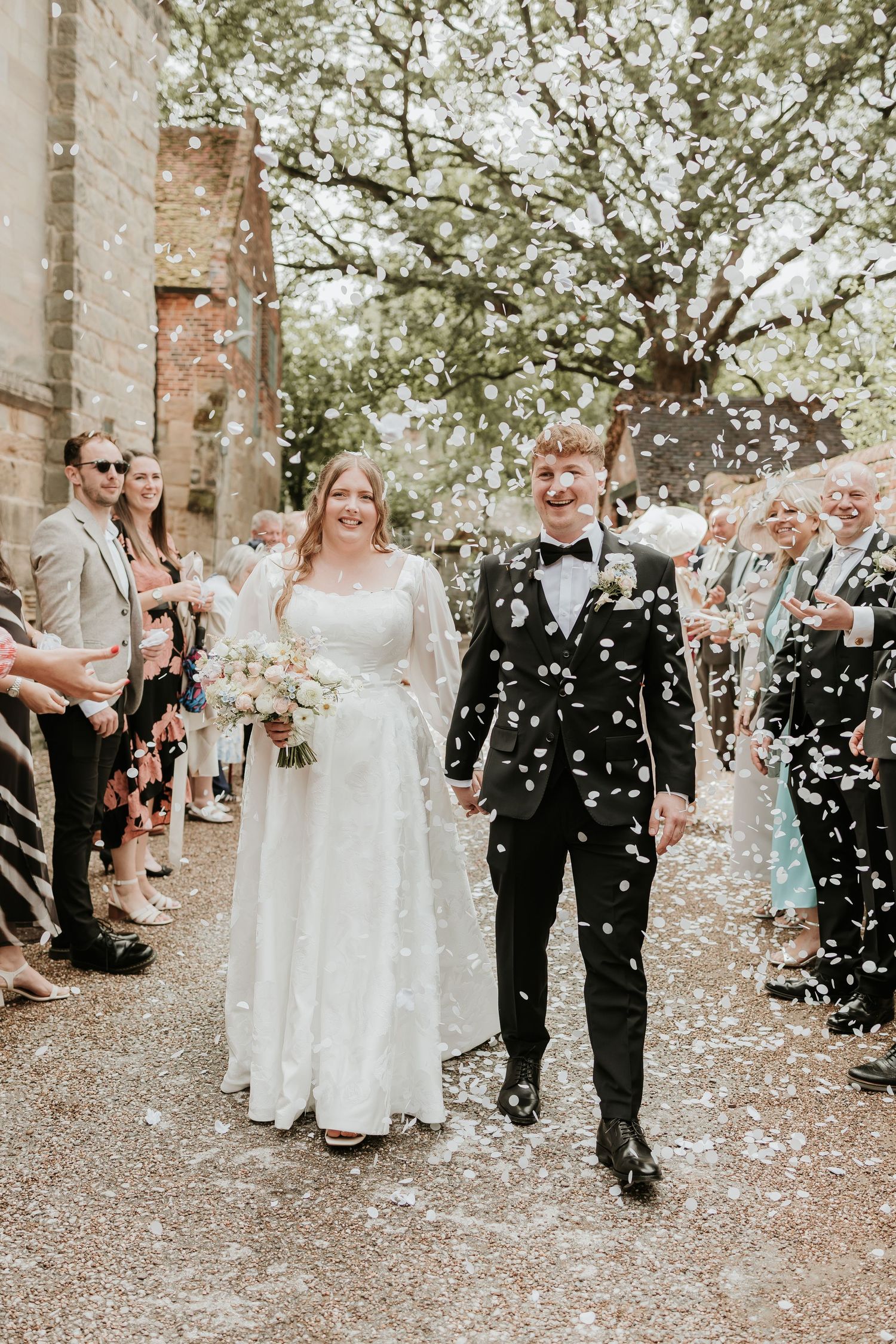 Newlyweds walk through confetti shower, bride in white gown holding bouquet, groom in black tuxedo, guests lining path.