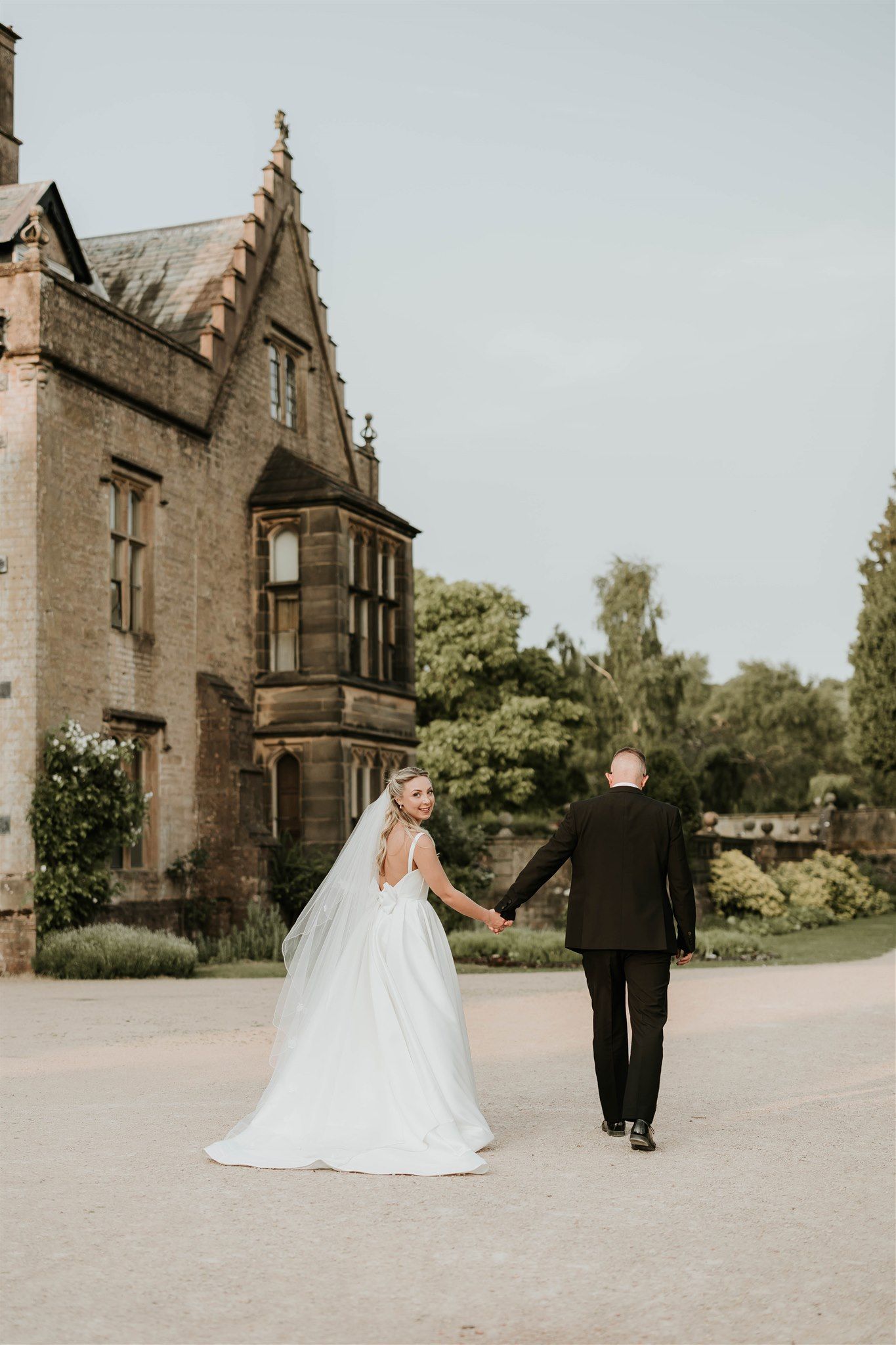 A couple walks hand in hand outside a Gothic-style stone manor house on their wedding day.