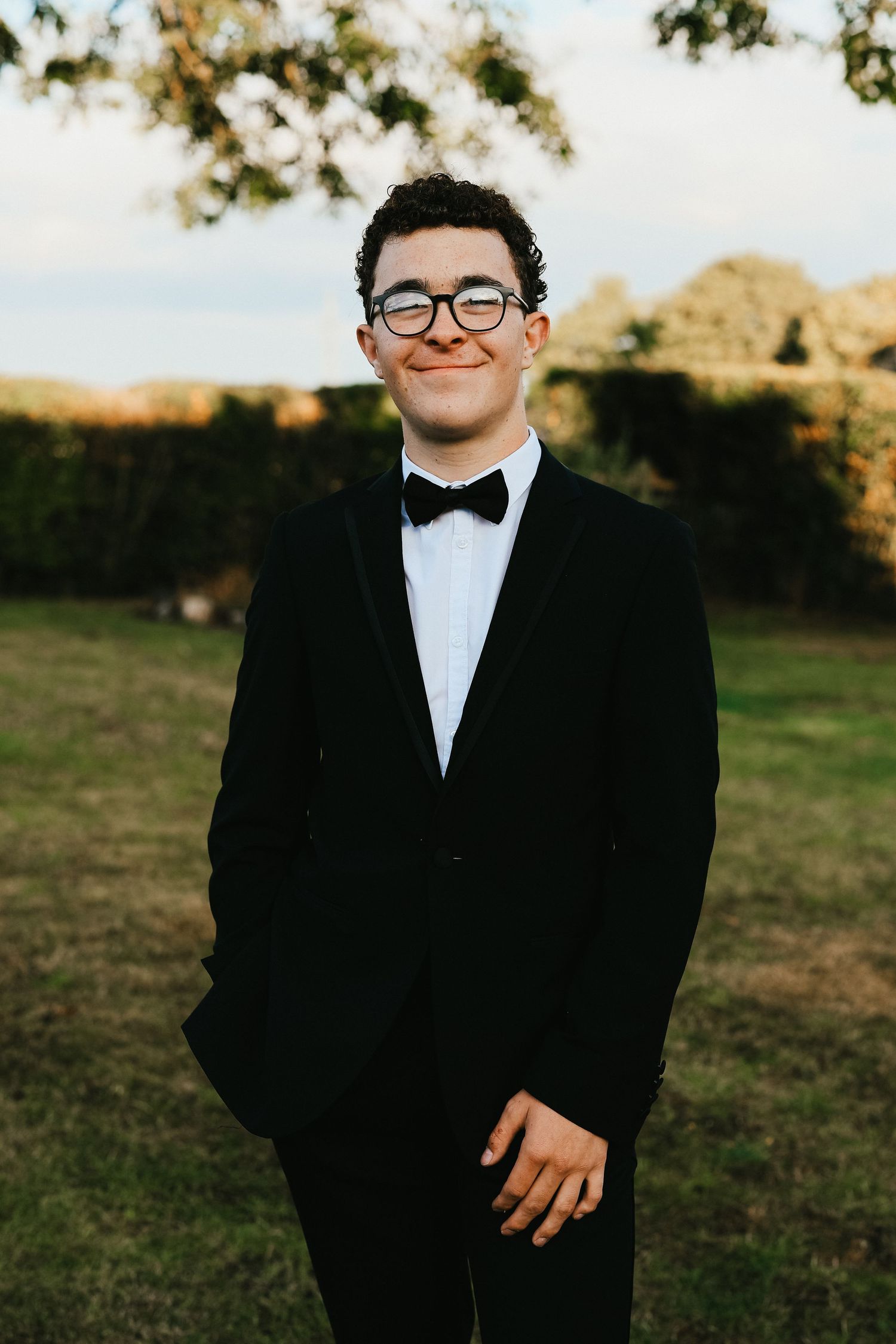 A person wearing a black tuxedo and bow tie smiles warmly while standing in a grassy field at golden hour.