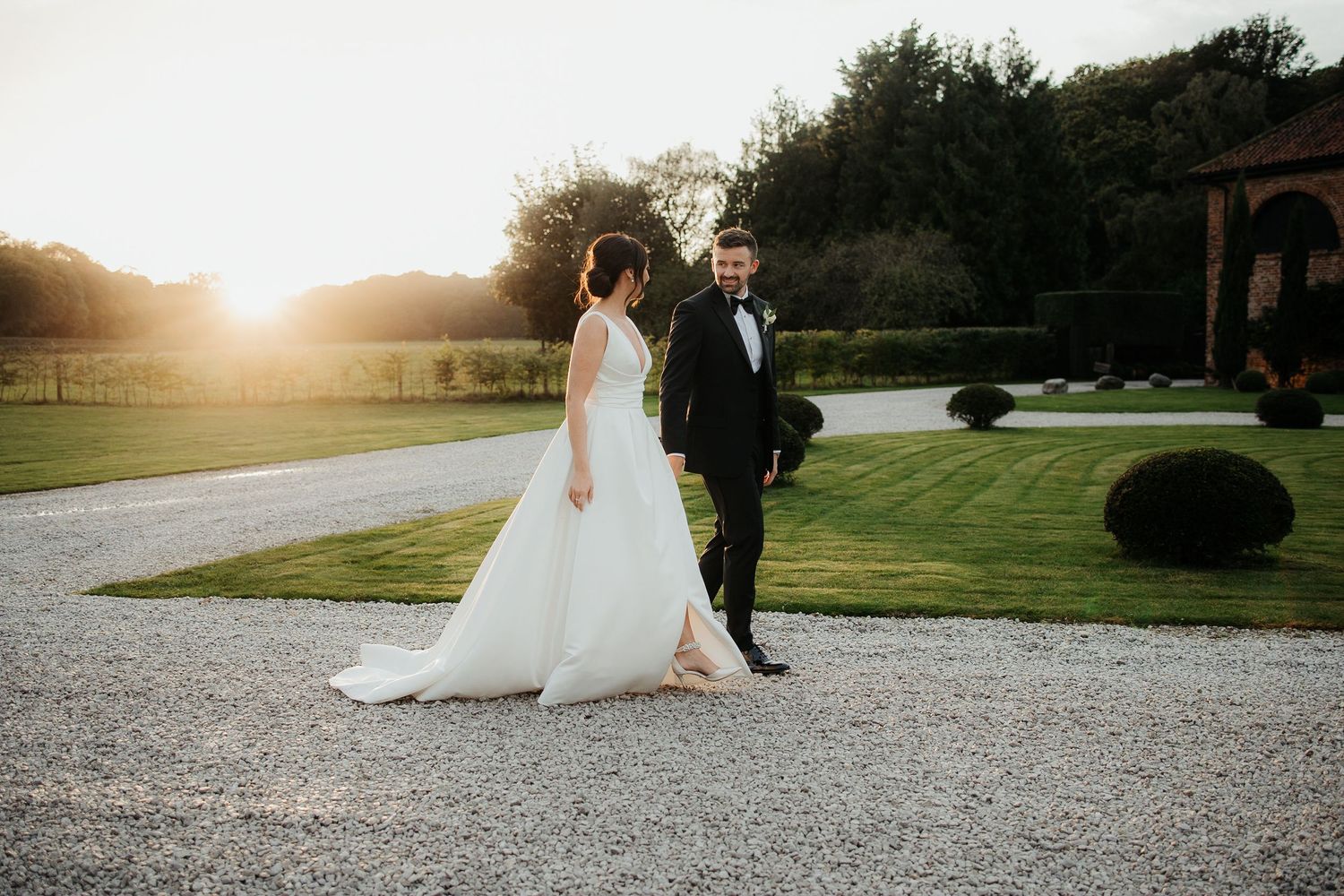 Bride and groom walking hand in hand on gravel path at sunset in elegant wedding attire at a countryside venue.