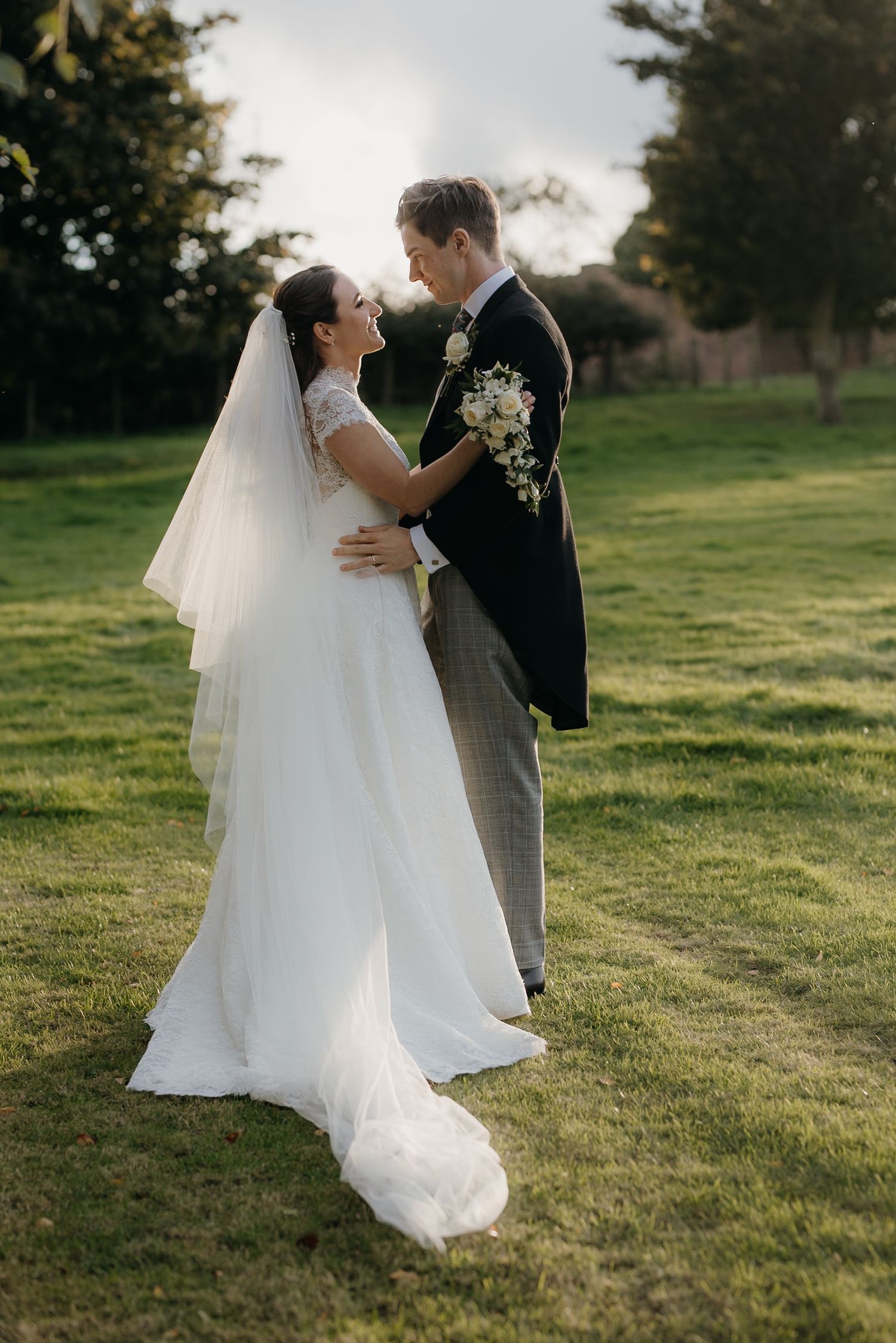 Bride in white gown and veil embraces groom in dark suit on lush green lawn during golden hour wedding portrait.