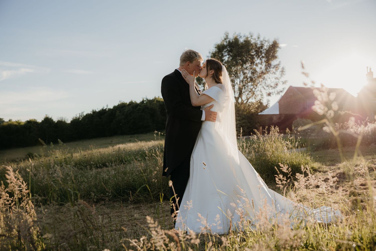 Bride and groom share a romantic kiss at golden hour in a sunlit field with a red barn in the background.
