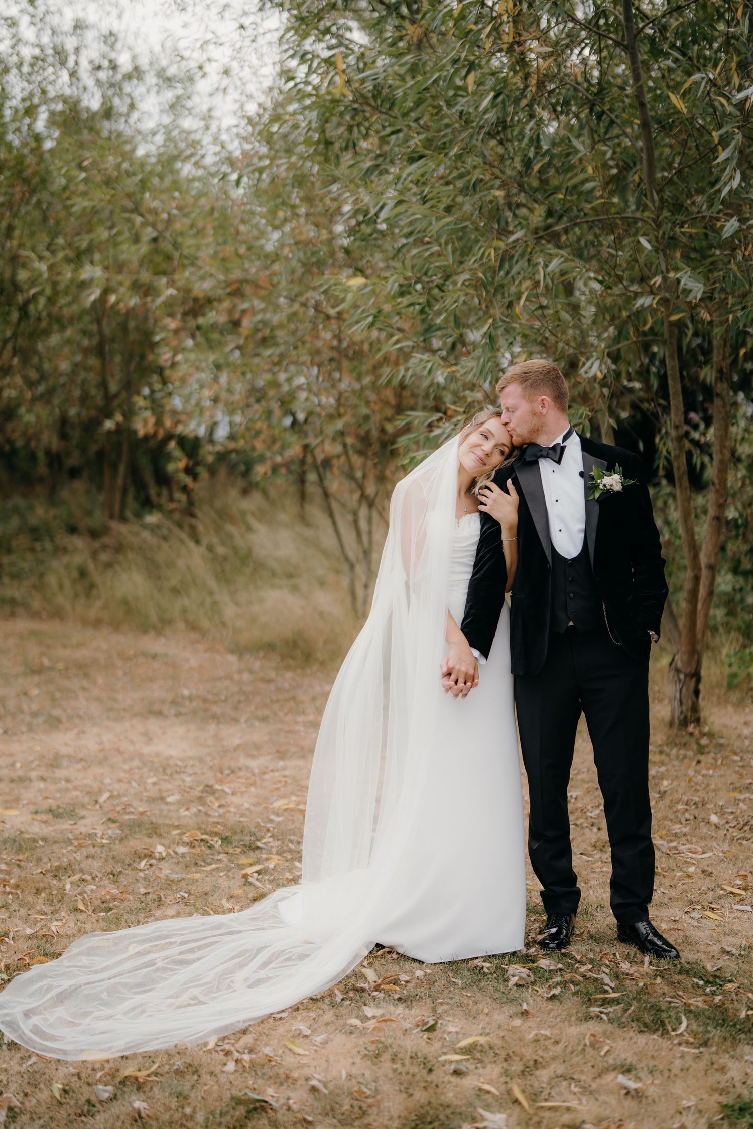 Bride in a long white gown and veil embraces a groom in a black tuxedo as he kisses her forehead among trees in an outdoor wedding portrait.