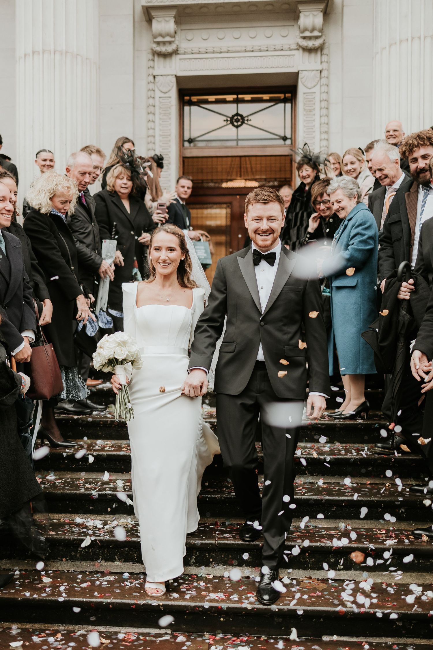 A bride and groom descend stone steps while guests shower them with confetti outside a historic building entrance.