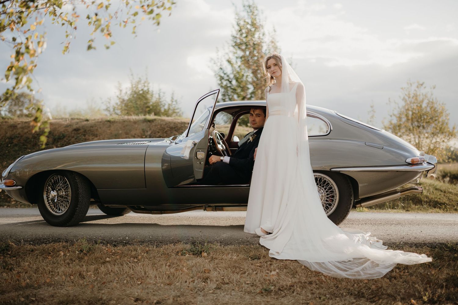 Bride in a long white wedding gown and veil stands beside a vintage silver Jaguar E-Type with the groom in a suit seated inside.