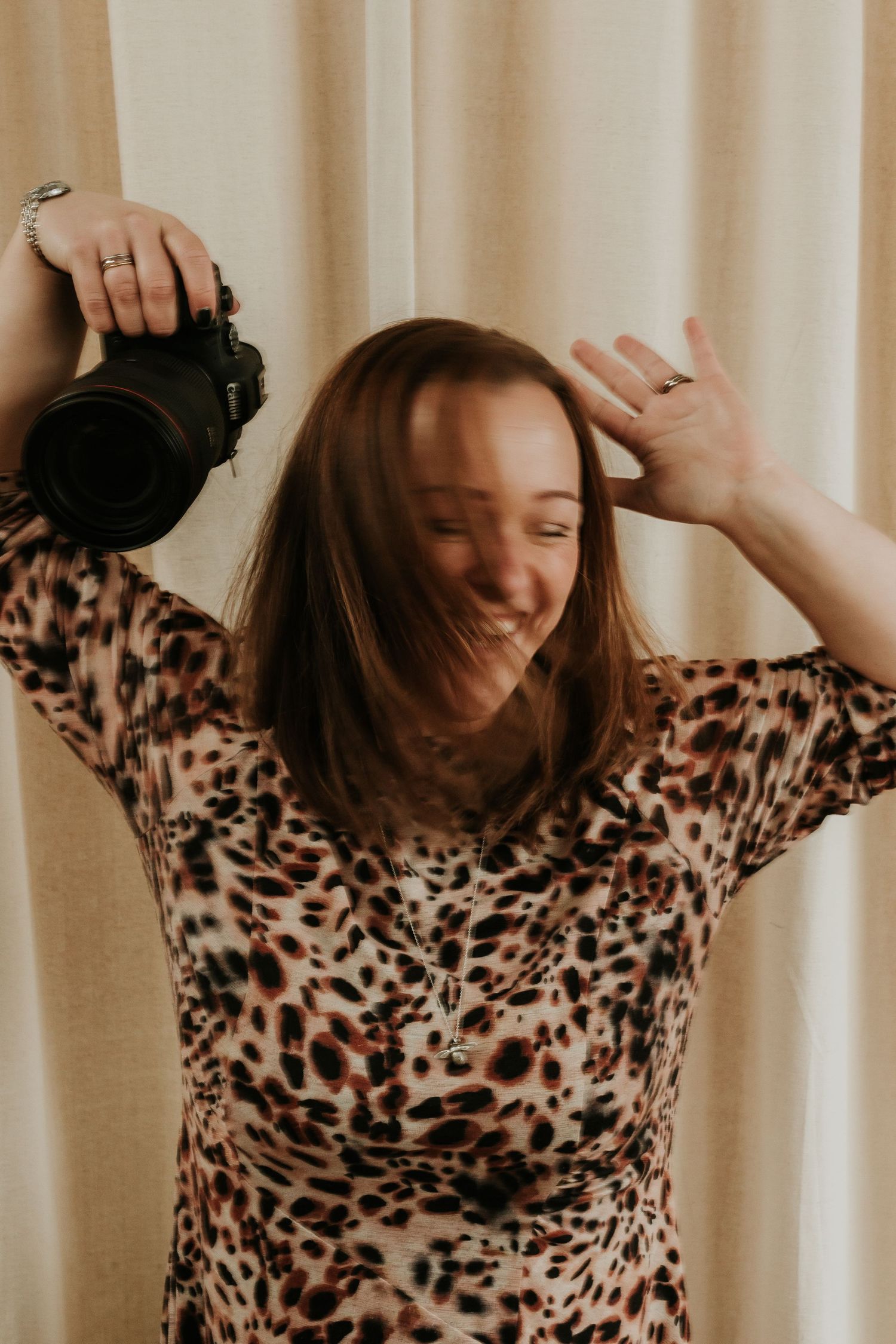 A laughing Sophie Ruck in a leopard print dress holds a DSLR camera while posing playfully against a cream curtain backdrop.