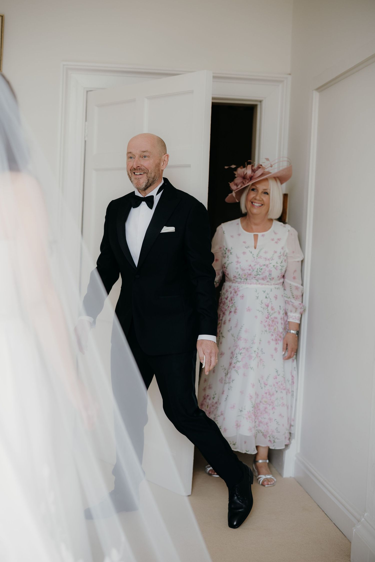 Smiling man in a black tuxedo and bow tie walks beside a woman in a floral dress and pink fascinator toward a bride in a veil.