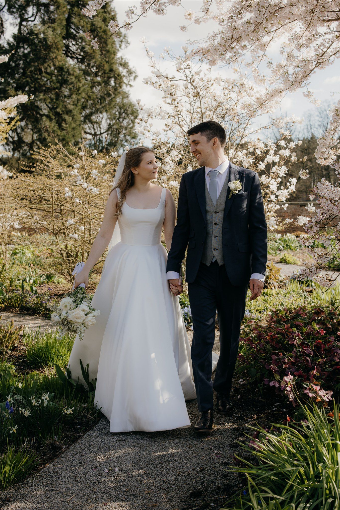 A bride and groom walk hand-in-hand through a garden path surrounded by blooming cherry blossoms on their wedding day at Middleton lodge.