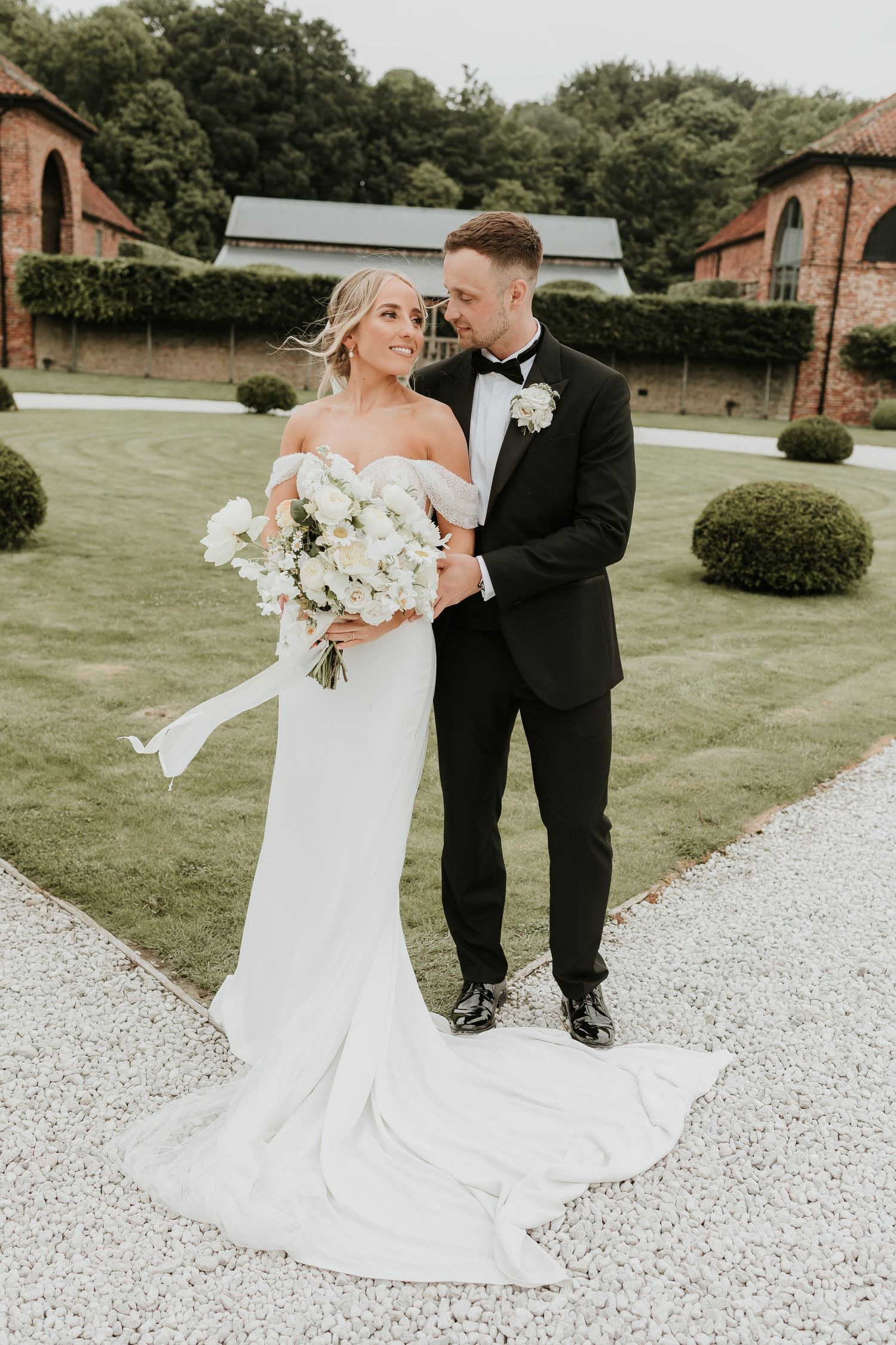 Newlyweds share a romantic moment on a manicured garden path with brick buildings and greenery in background of Hazelgap barn.